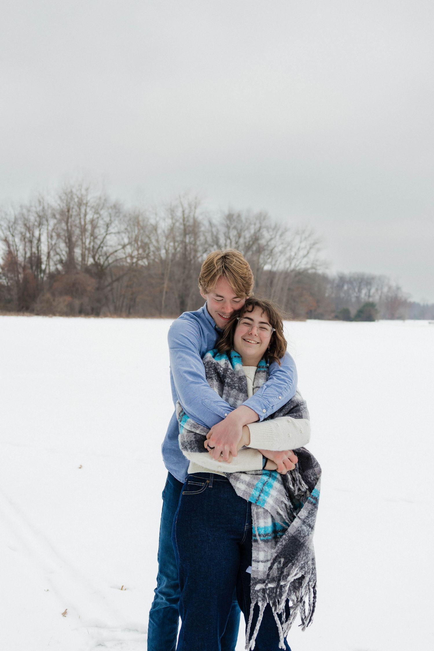 Couple embracing warmly under a plaid blanket in a snowy open field engagement photo.