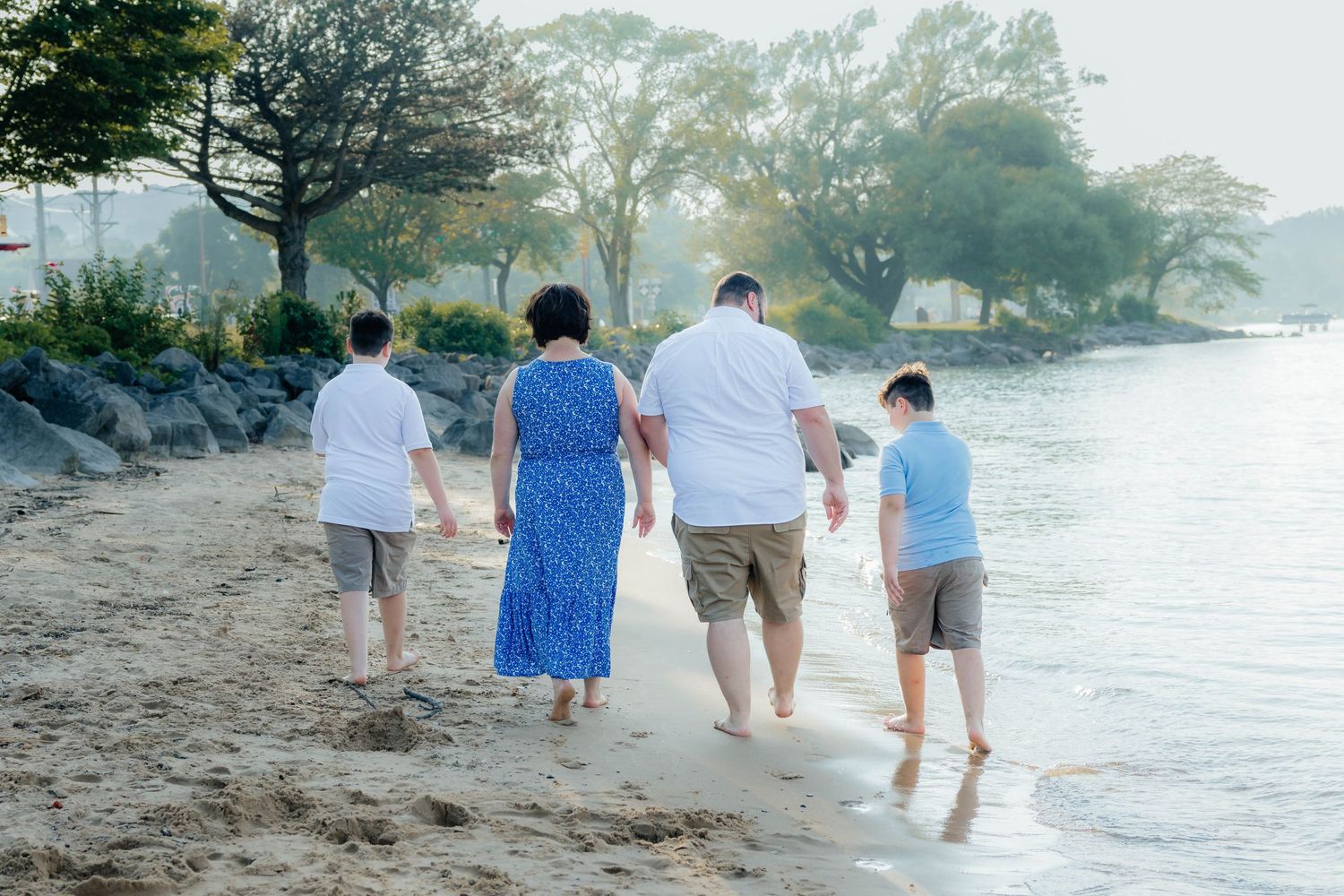 Family of four walking barefoot along a sandy beach near the water's edge.