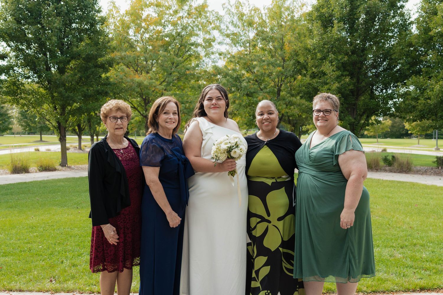 Bride in white gown poses with four women in formal attire on green lawn outdoors.