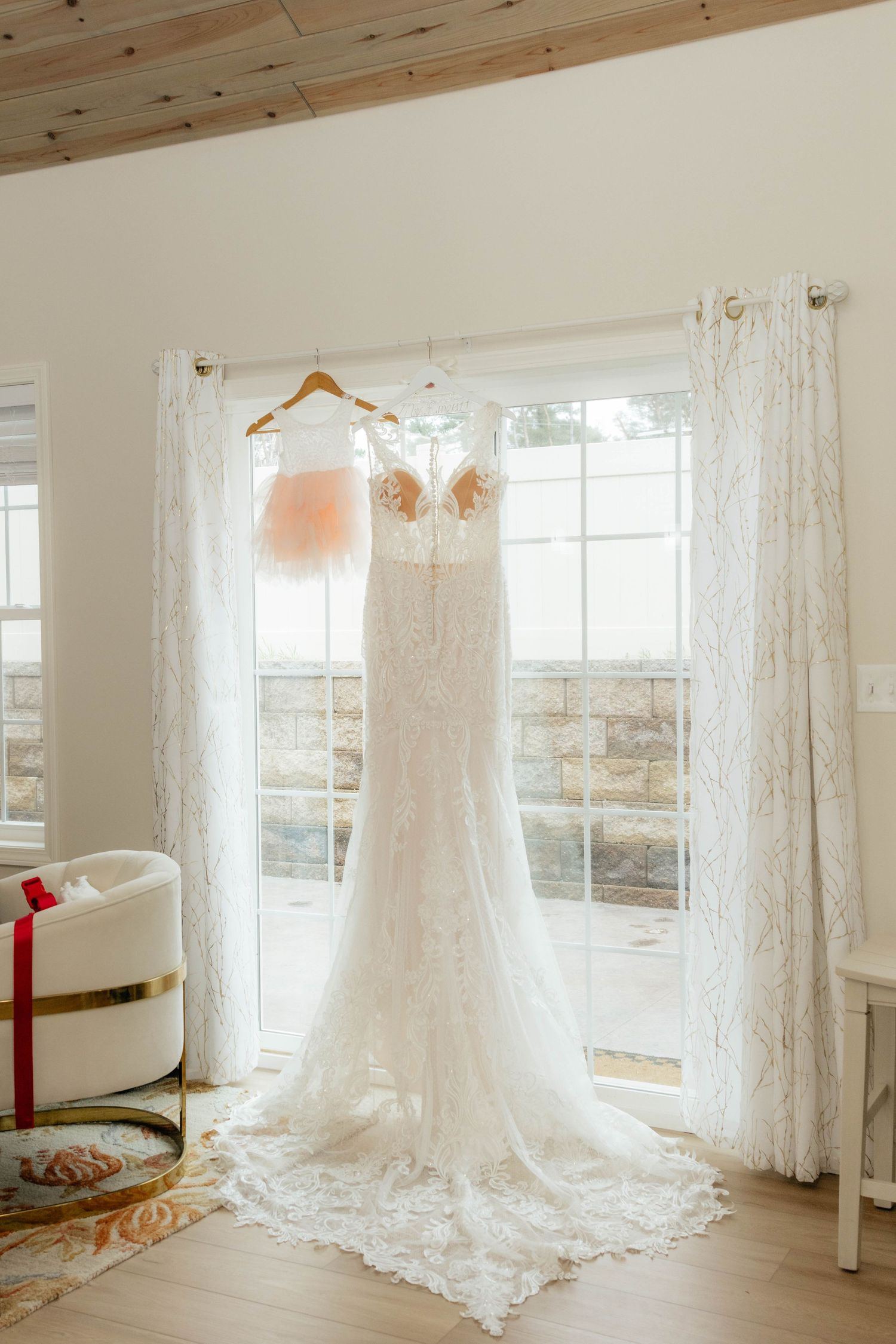 Elegant lace wedding dress and blush bridesmaid gown hanging on a wooden hanger in a bright window.
