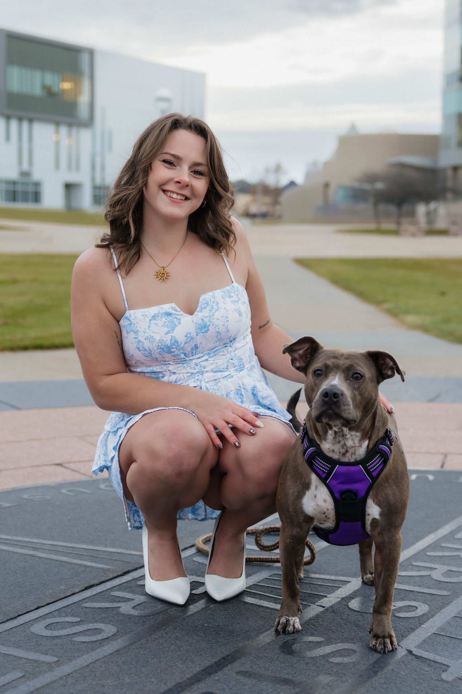 Smiling young woman in blue floral dress crouches beside her brown pit bull dog wearing a purple harness on campus.