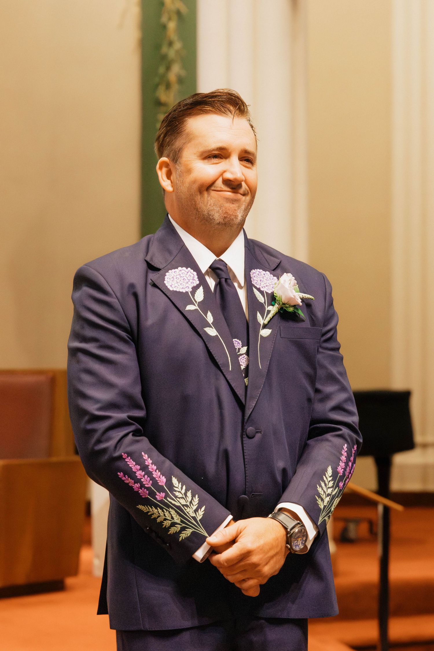 Emotional groom in navy embroidered suit smiles with tears at church altar during wedding ceremony.