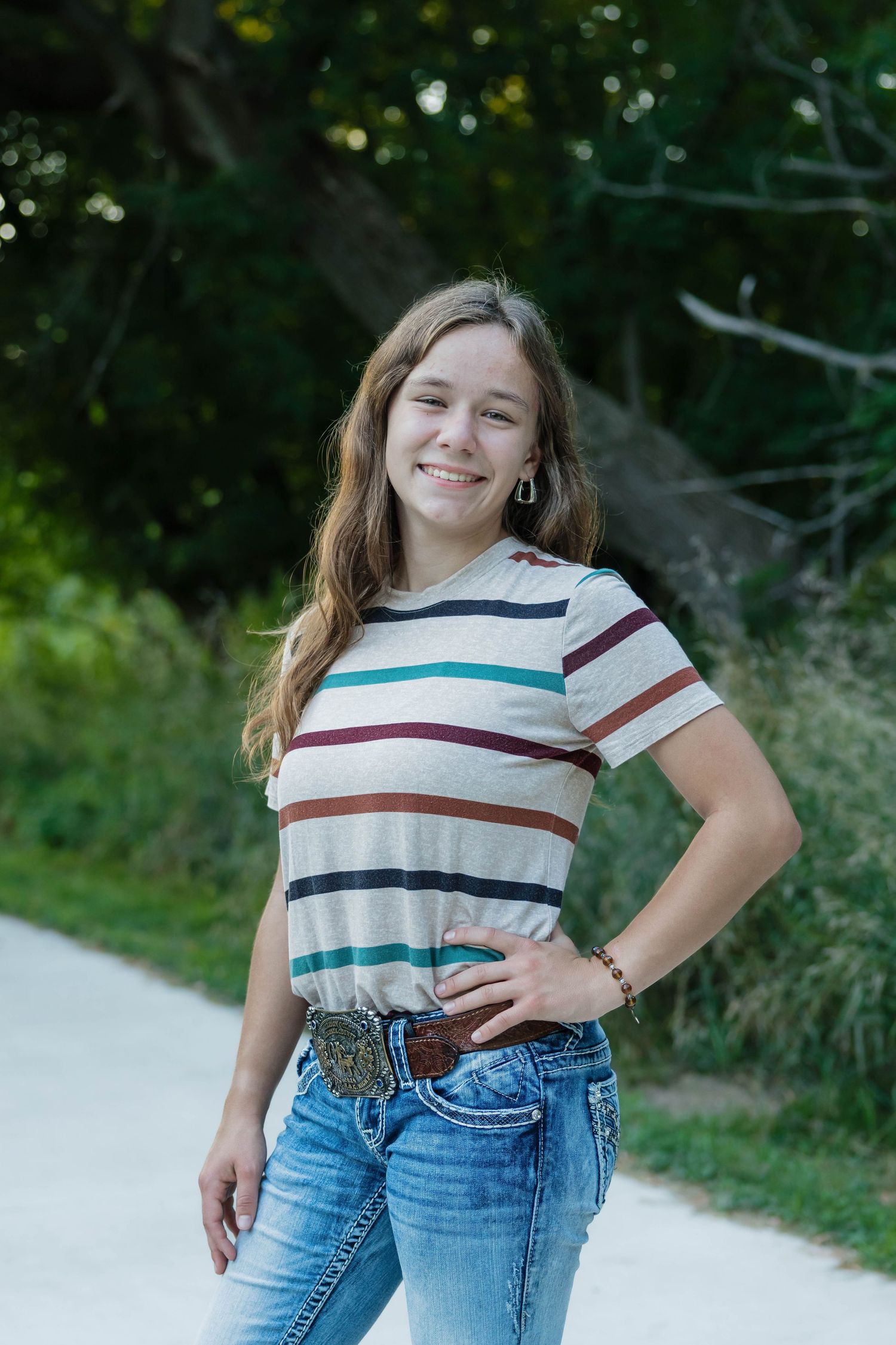 Smiling teenage girl in striped shirt and jeans with a decorative belt posing outdoors on a path.