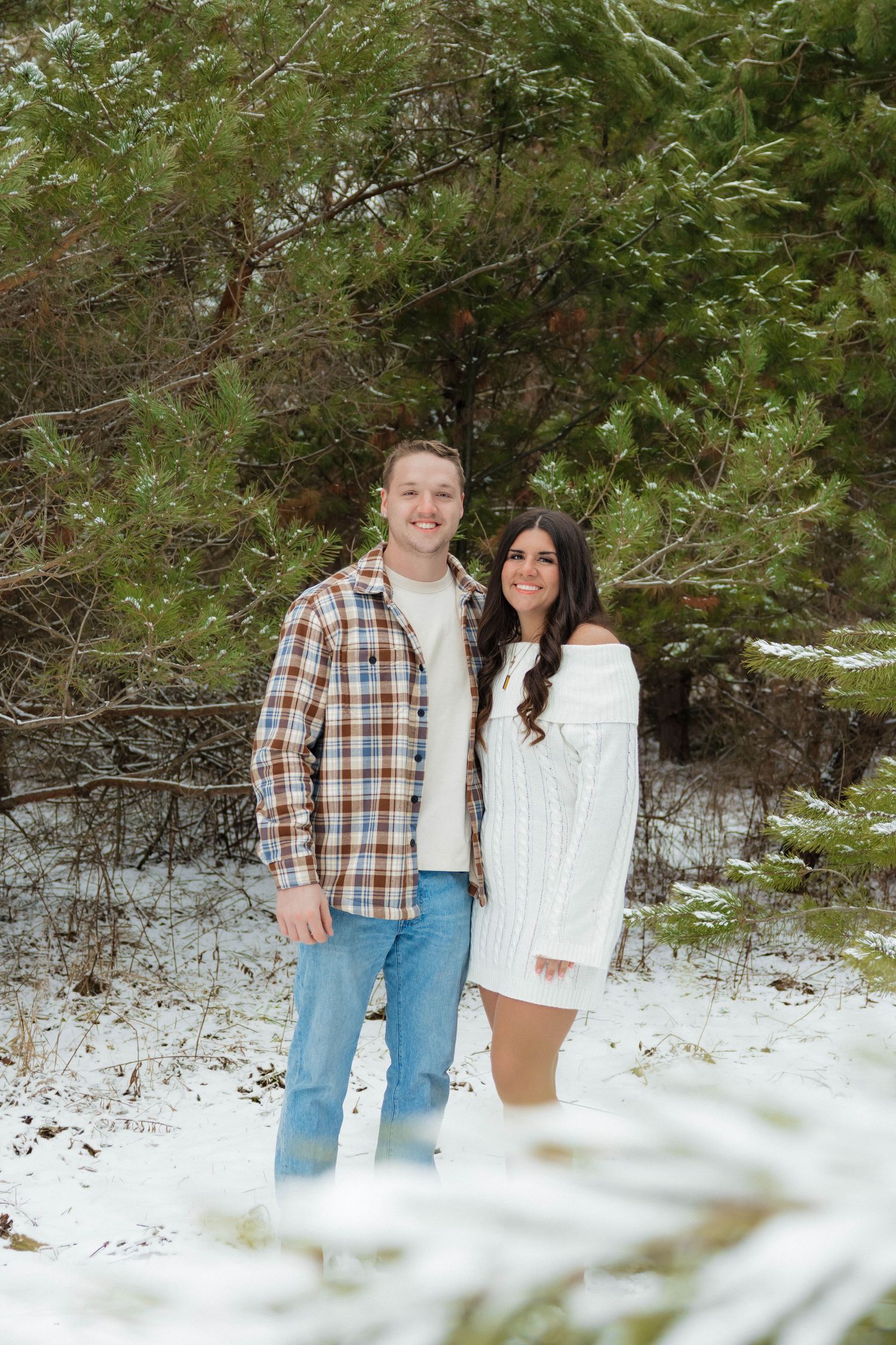 Smiling couple standing together in a snowy forest, man in plaid shirt and woman in white knit dress.