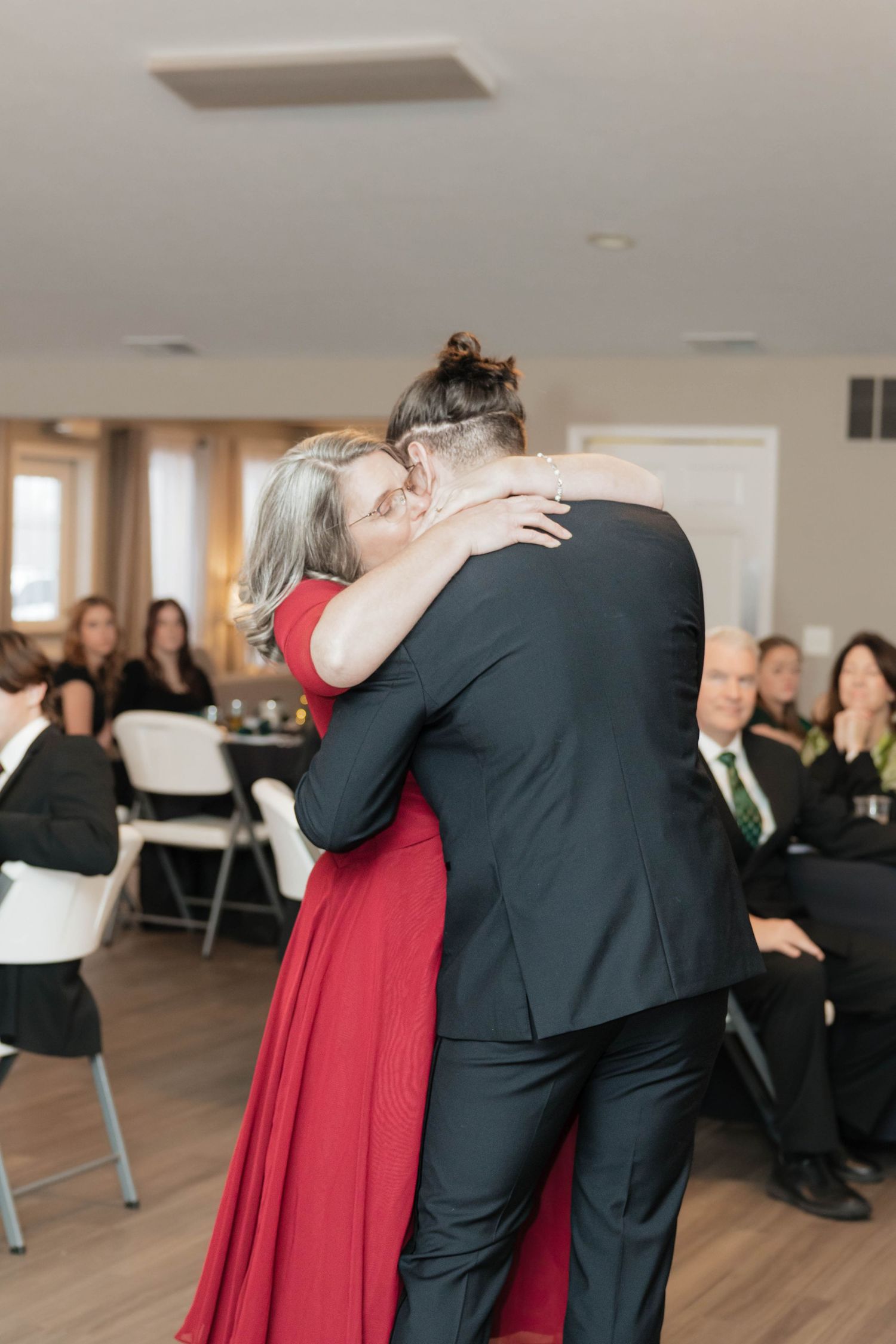 Mother and groom share emotional dance embrace at wedding reception with guests seated nearby.
