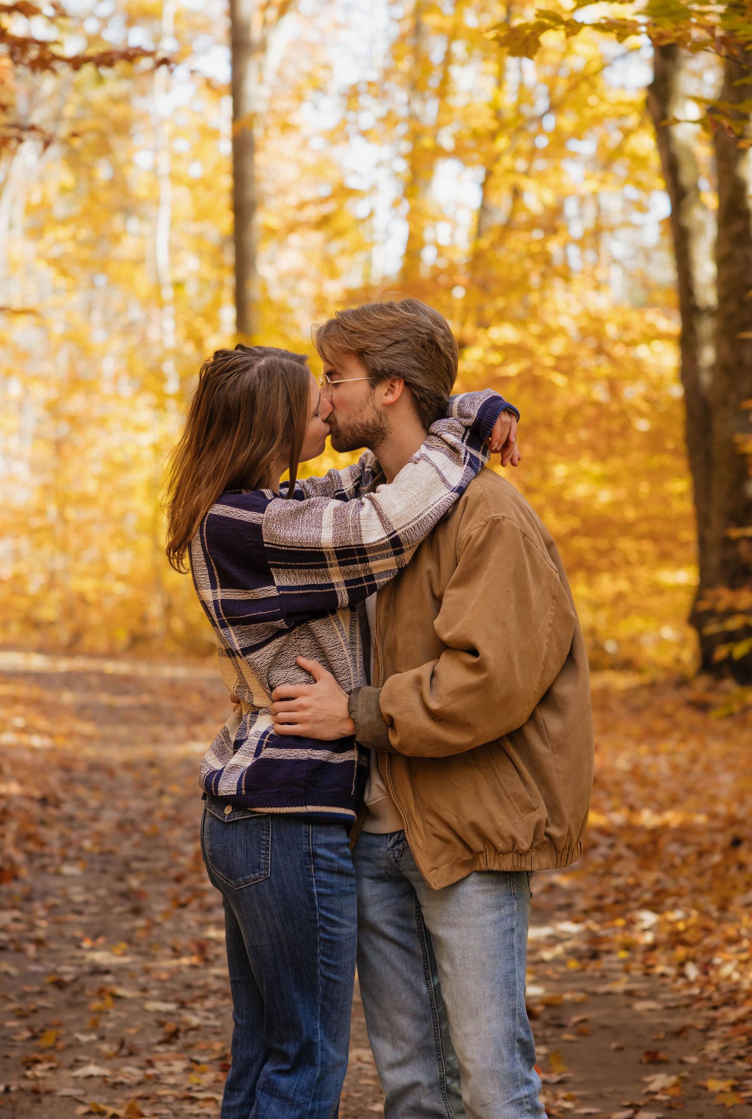 Couple embracing and kissing on a leaf-covered trail surrounded by vibrant golden autumn trees.