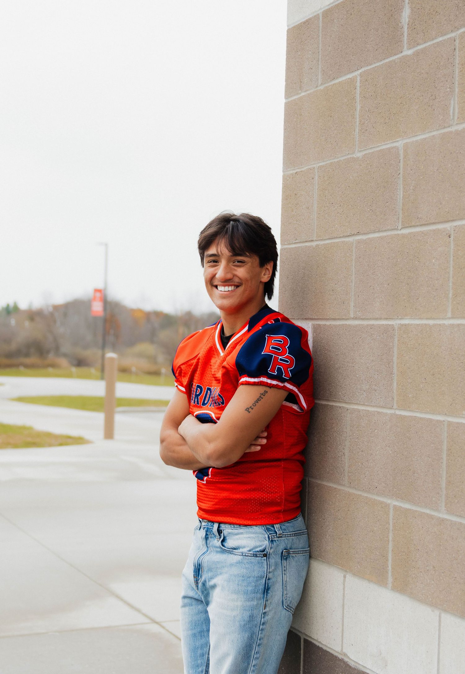 Smiling teen in orange BR football jersey leans against brick wall with arms crossed.