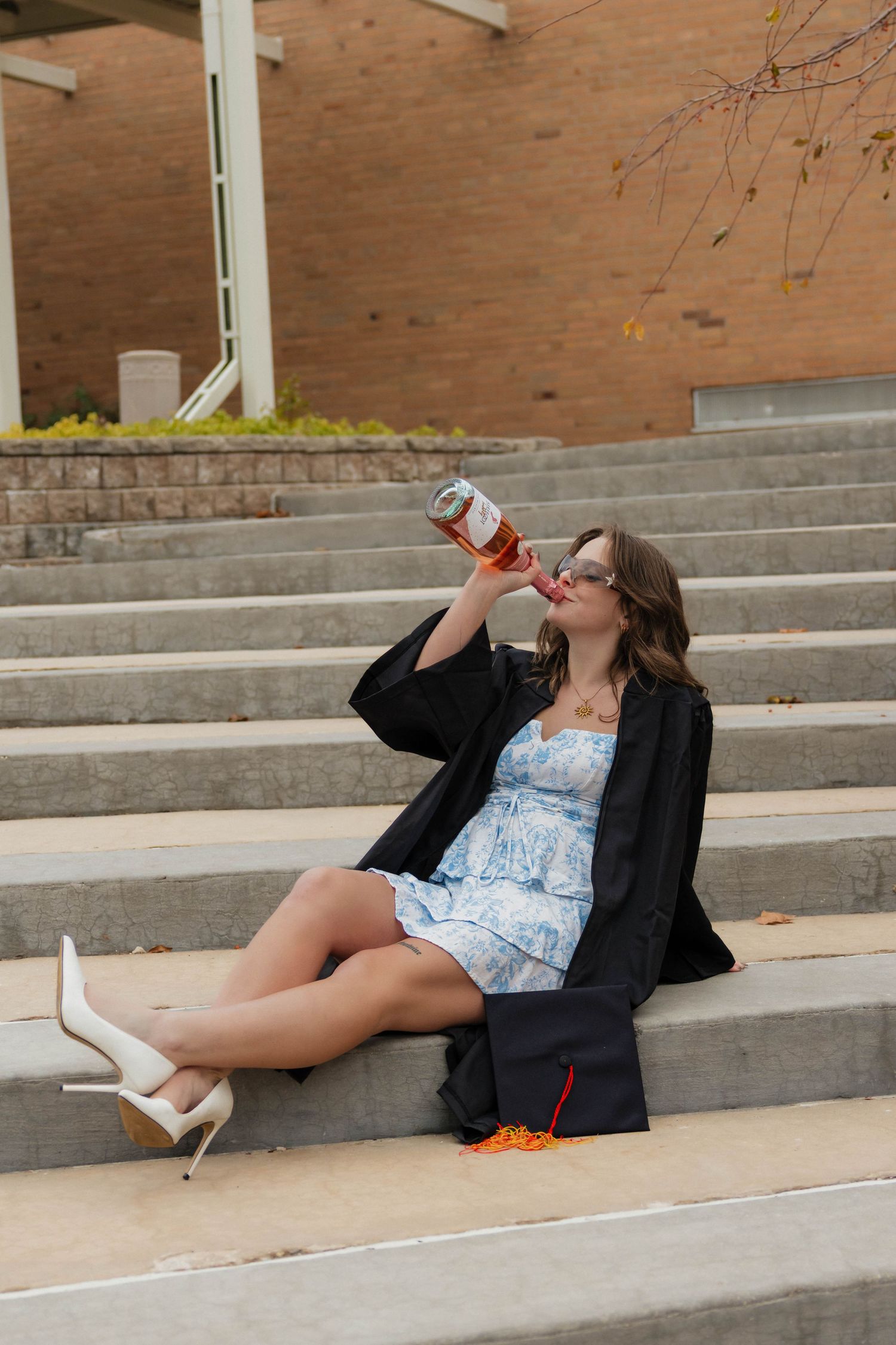 Female graduate in cap and gown sitting on outdoor steps drinking from a bottle on university campus.