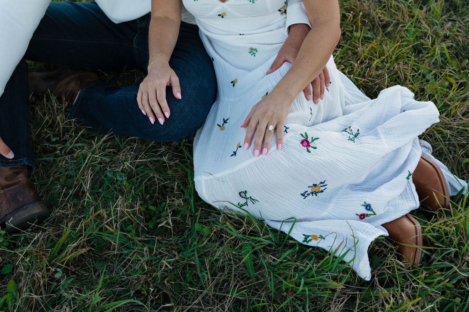 Close-up of an engaged couple's hands in grass, woman wearing a diamond ring and floral white dress.