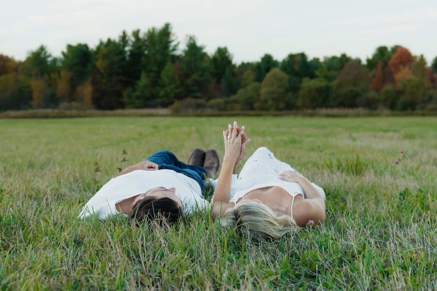 Romantic couple lying in a green meadow with a tree line visible in the background.