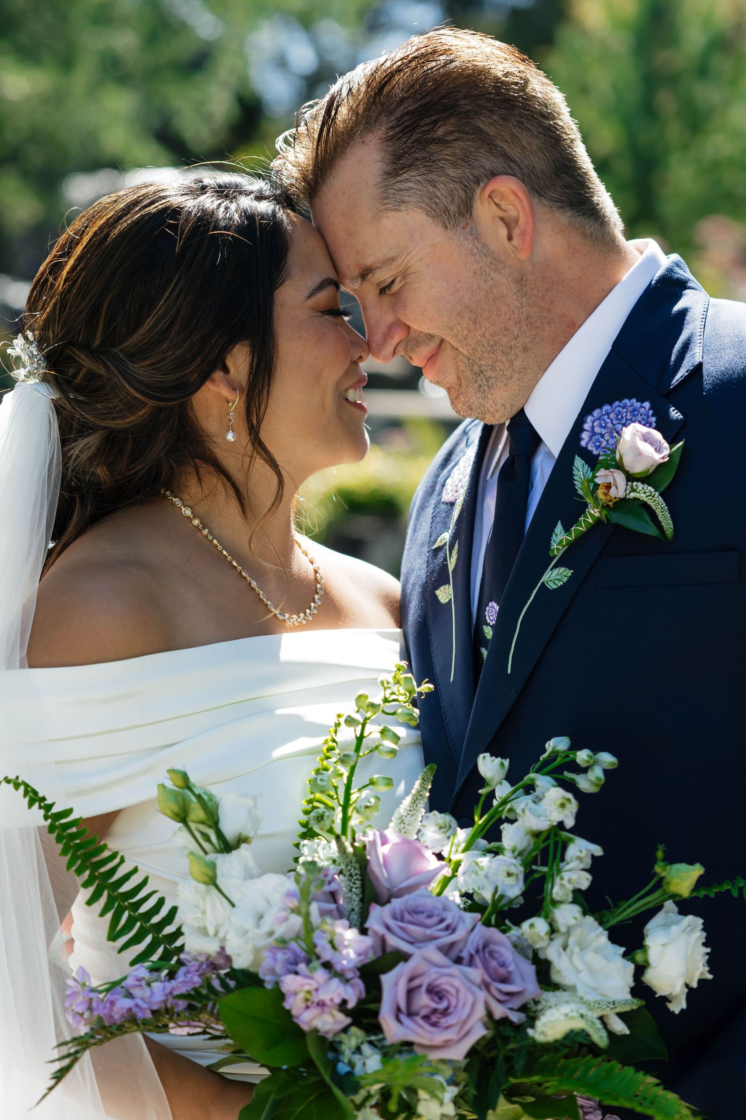 Bride and groom touching foreheads and smiling holding purple and white wedding bouquet outdoors.