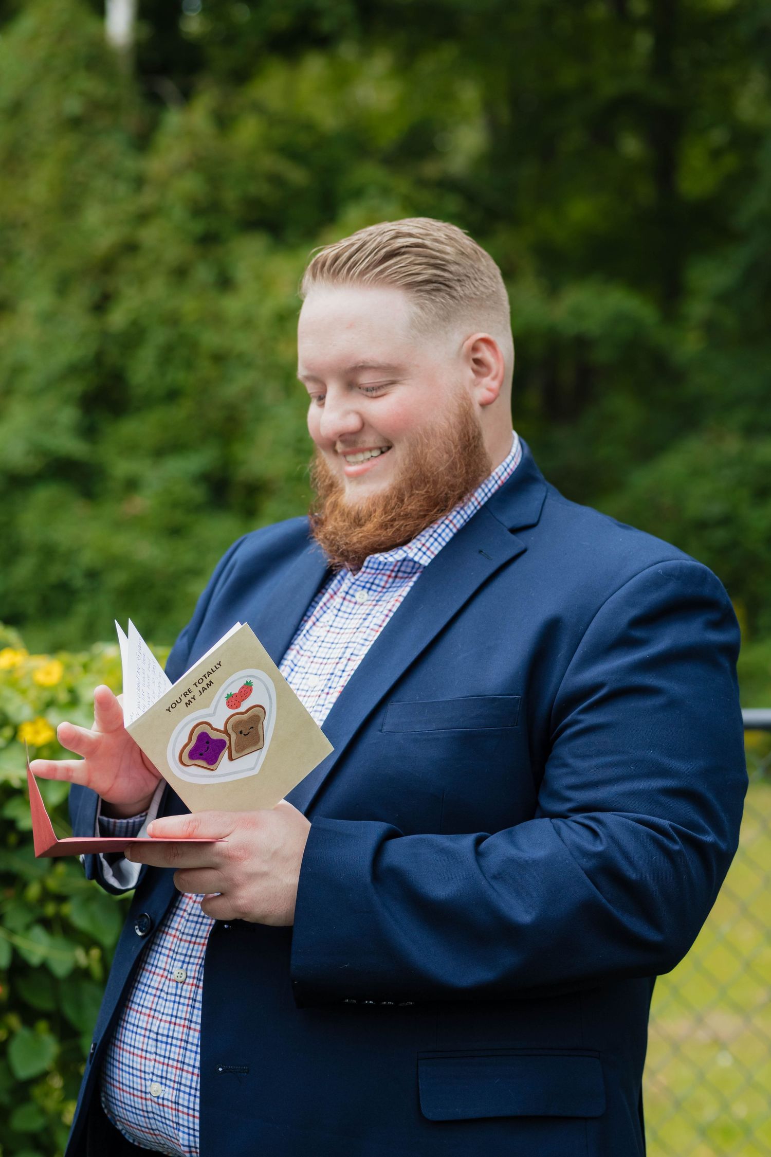 Smiling groom in navy suit reads a heartfelt card outdoors surrounded by lush greenery.