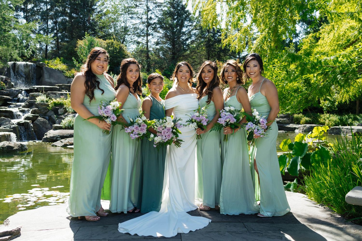 Bride with bridesmaids in sage green dresses holding pink and purple bouquets pose by garden waterfall.