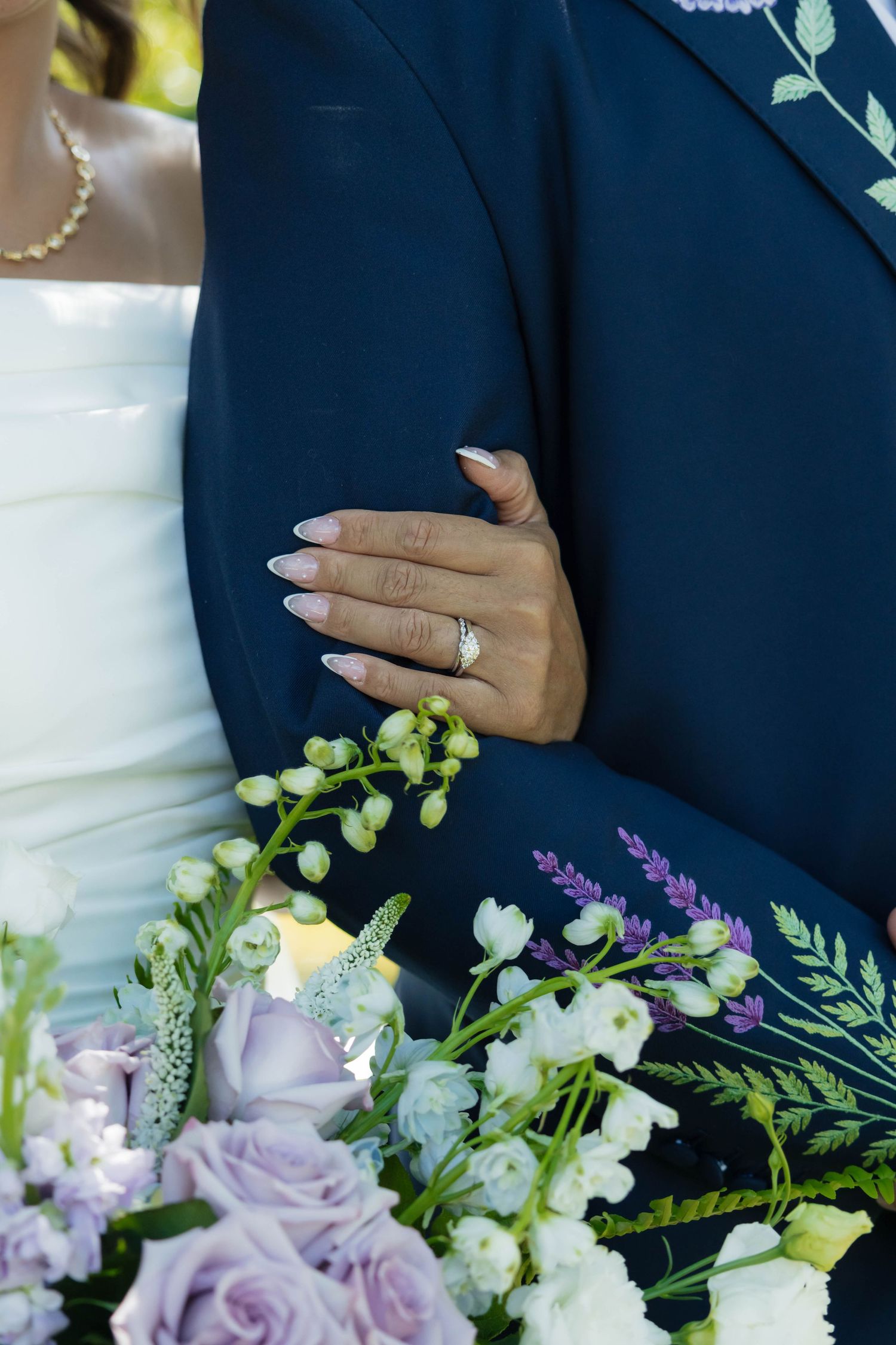 Close-up of bride's hand with diamond engagement ring resting on groom's navy suit jacket.
