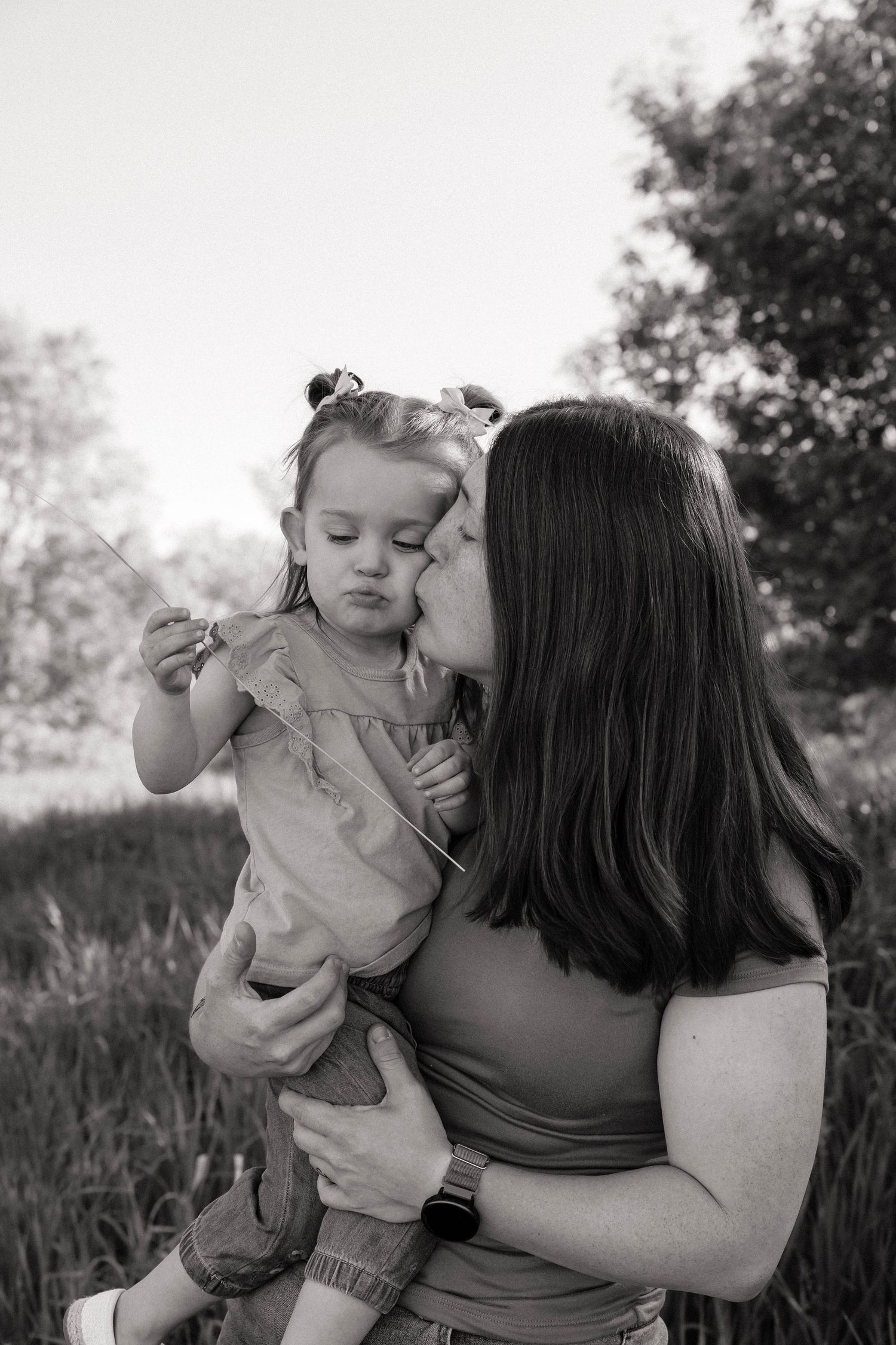 A mother kissing her toddler daughter on the cheek in a black and white outdoor portrait.