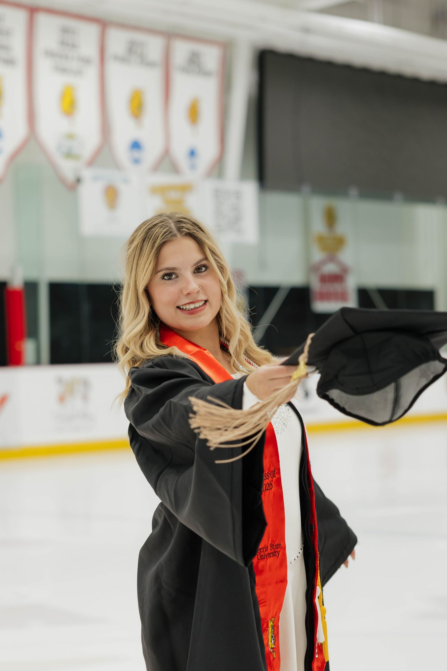 Female figure skater in graduation gown and orange stole poses on ice rink holding cap tassel.