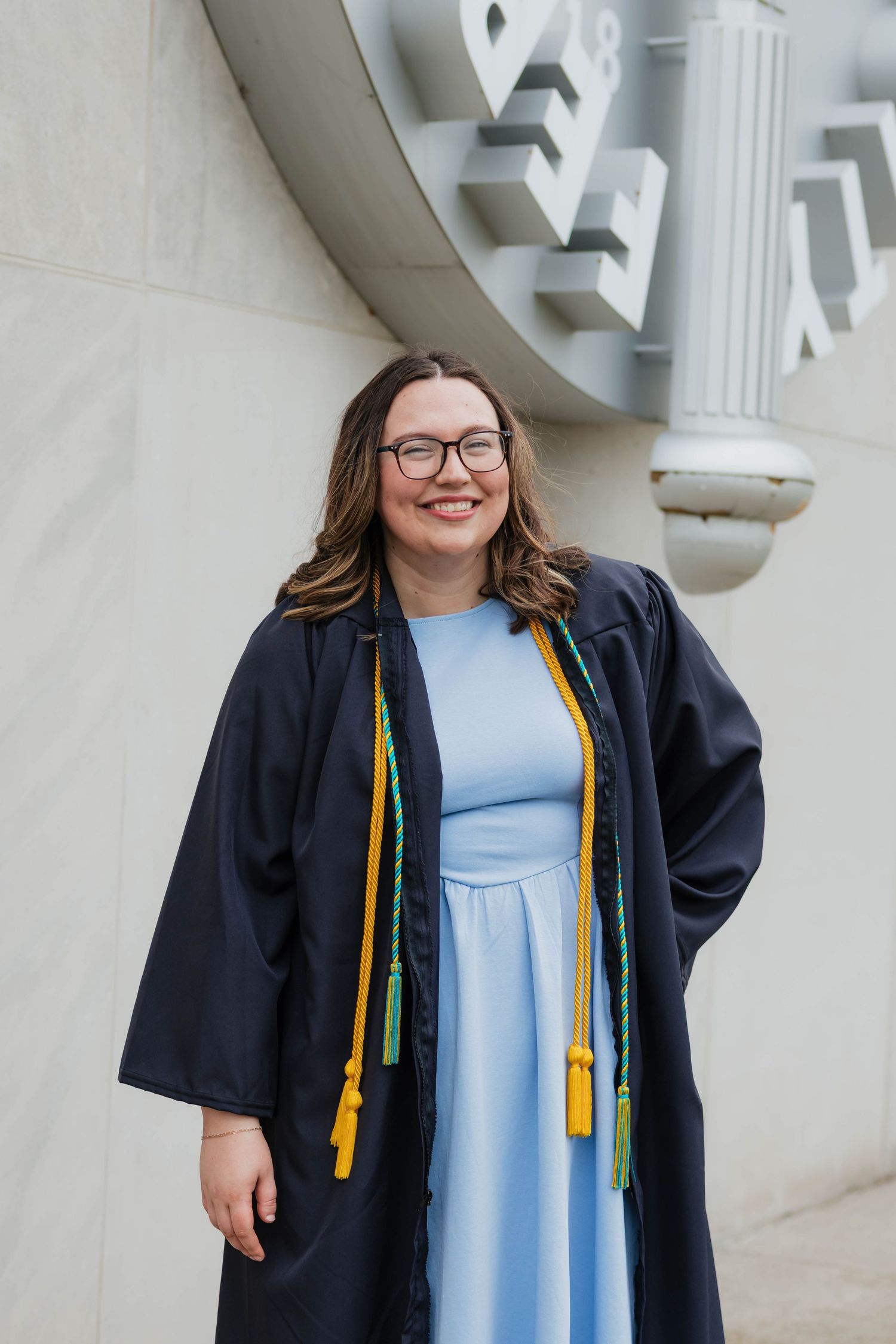 Smiling graduate in black gown with green and gold honor cords posing outside Ferris State University building.