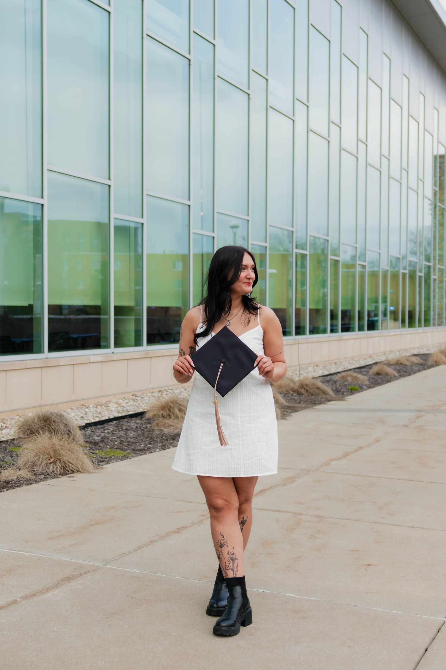 Brunette graduate in white dress holding black mortarboard cap outside a modern glass building.