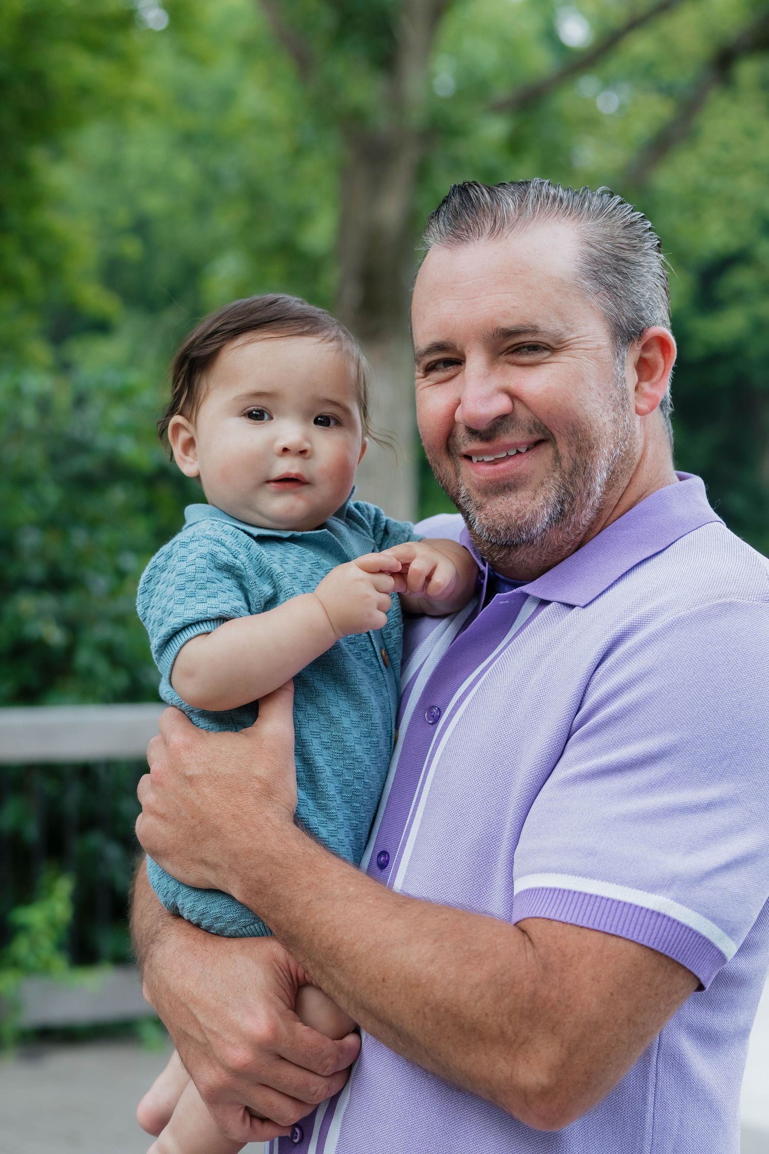 Proud father holding his baby boy while smiling outdoors in a park.