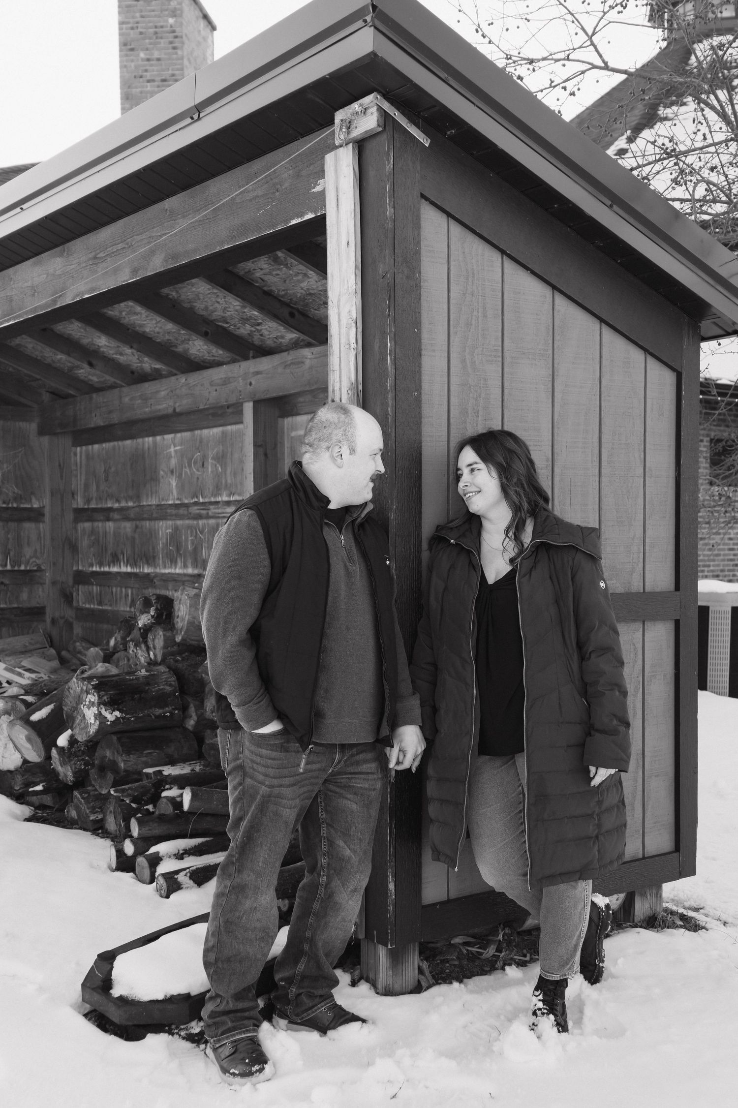 Black and white photo of a couple holding hands beside a wooden shed in snowy landscape.