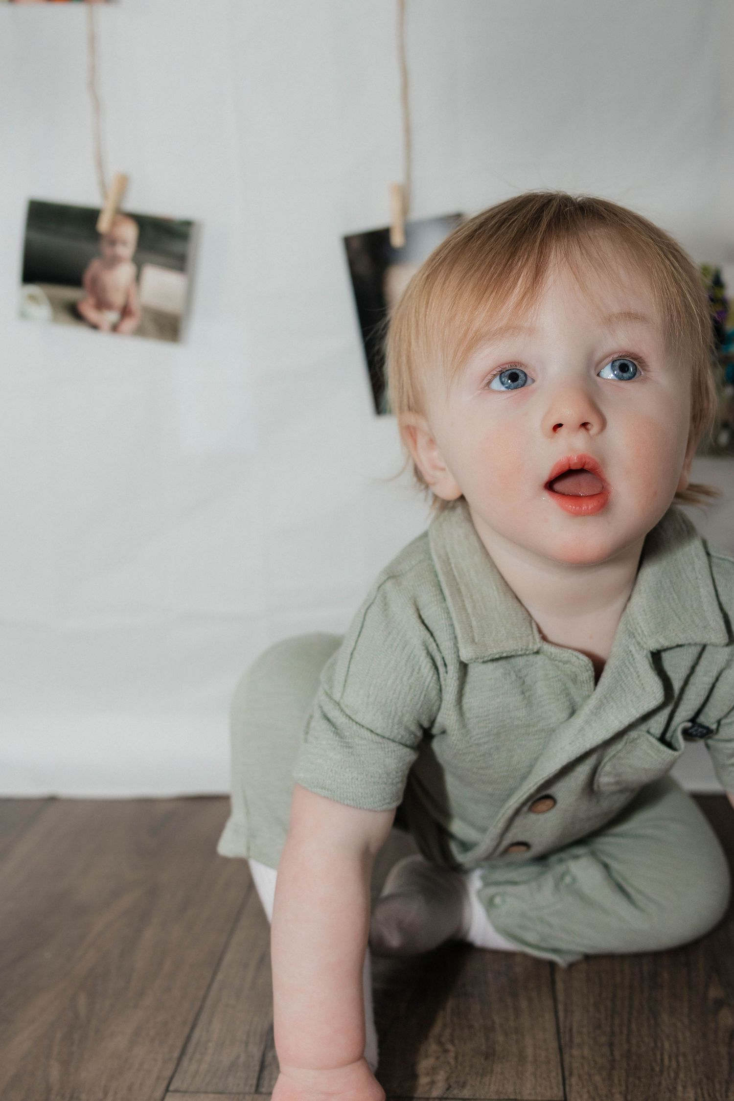 Blue-eyed toddler in sage green romper sits on wood floor at first birthday photo session.