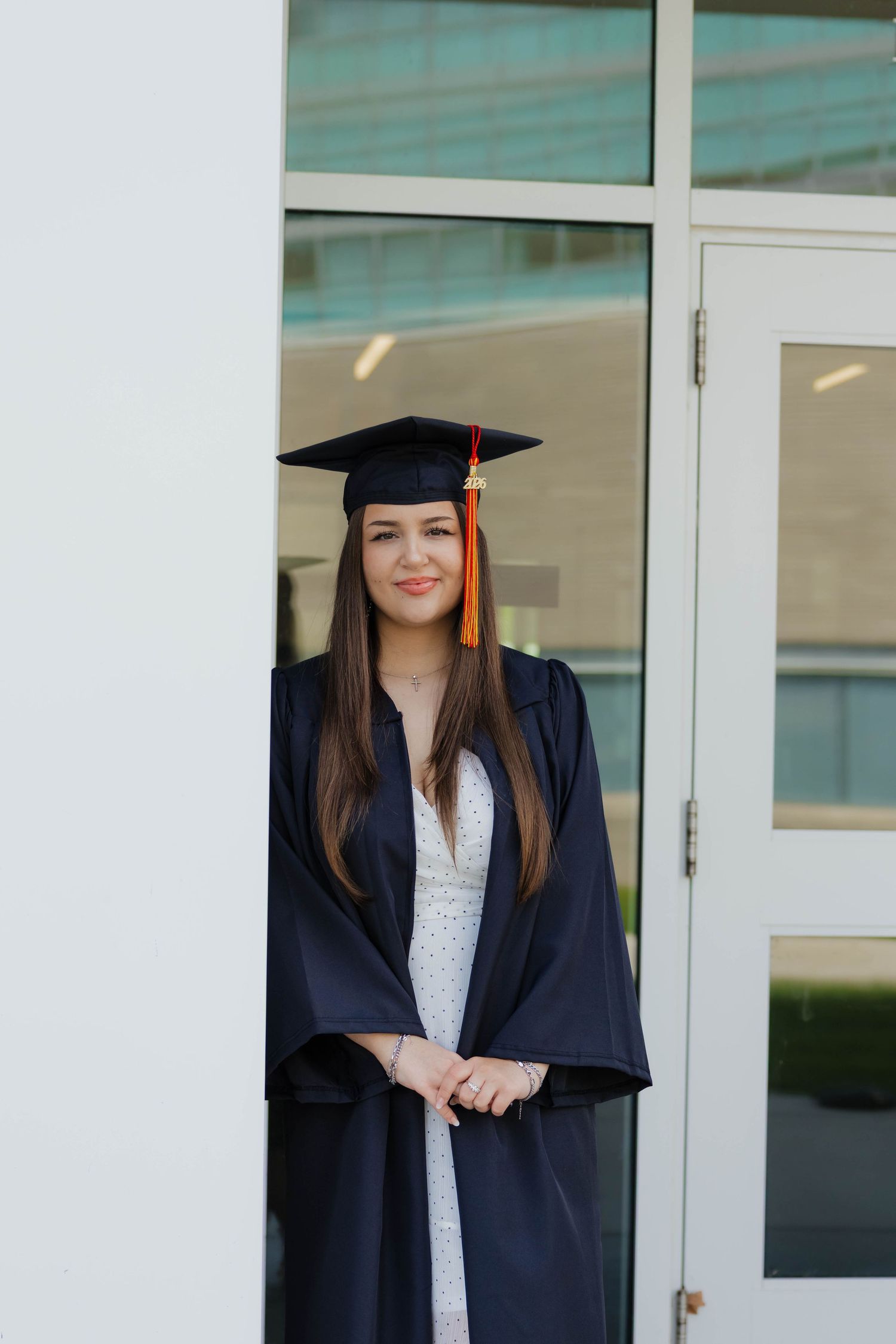 Young woman in navy graduation gown and cap with orange tassel poses by white building doors.
