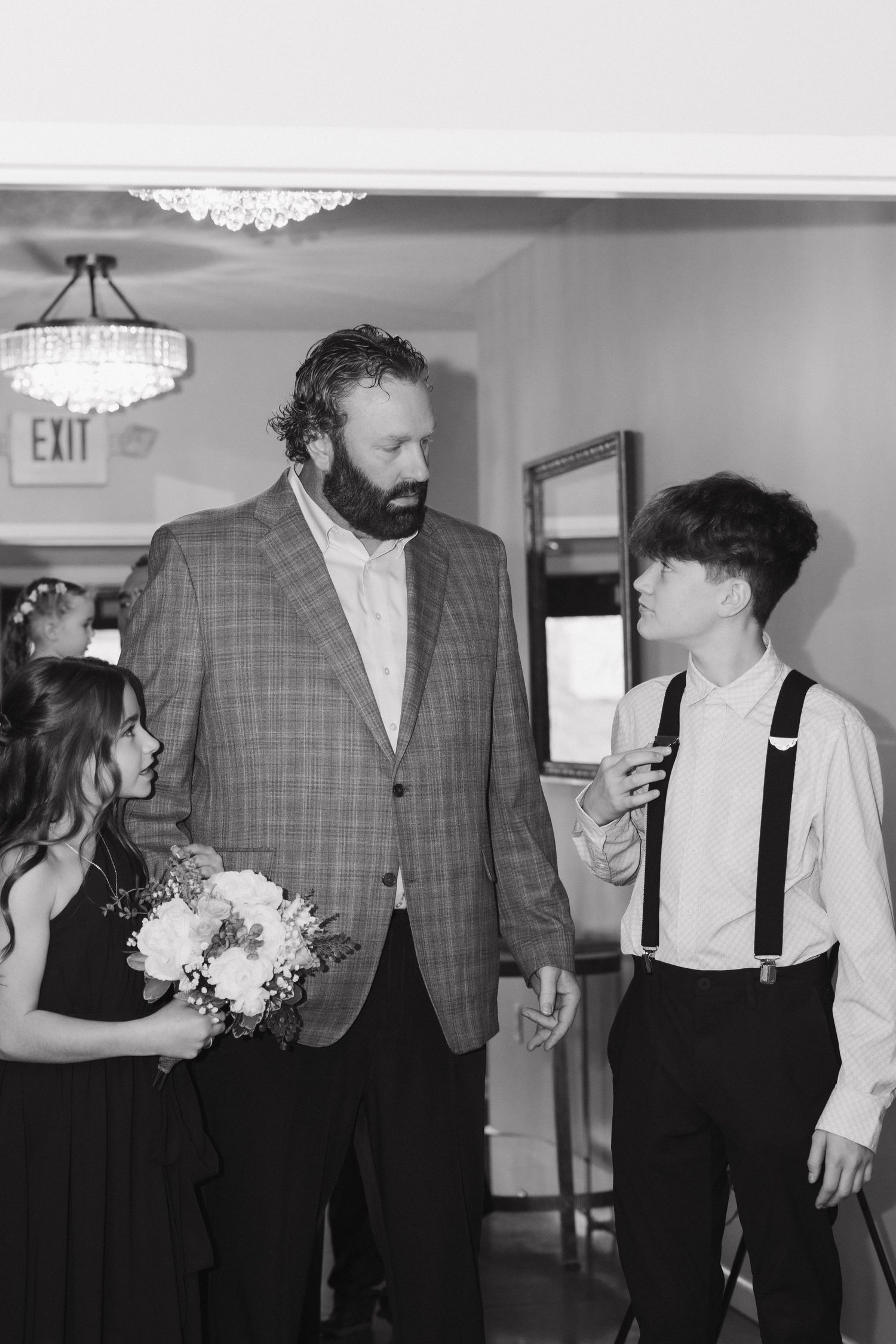 Black and white photo of bearded man in plaid suit speaking with children in wedding attire indoors.