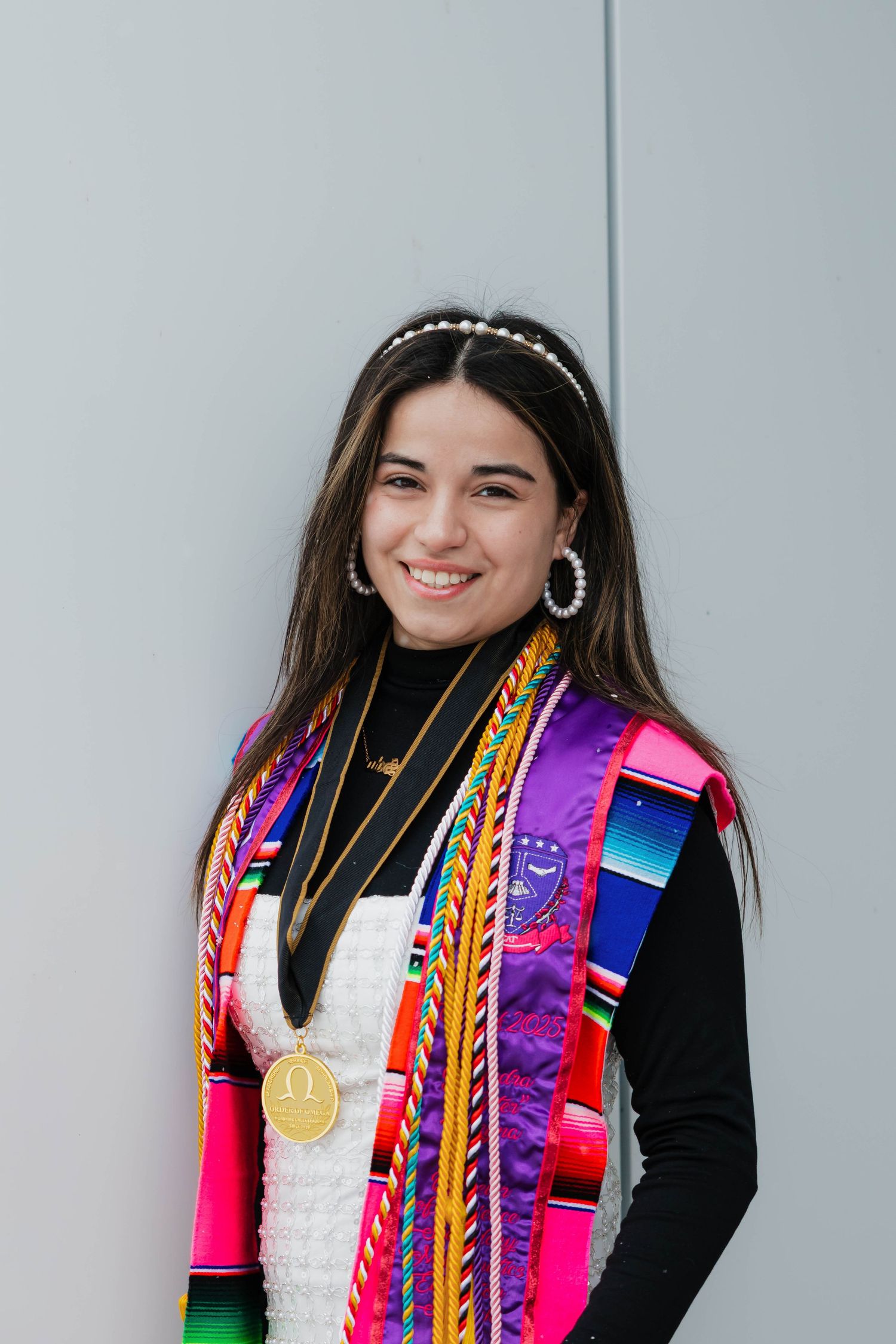 Smiling graduate wearing colorful serape stole and gold medal against a gray wall backdrop.