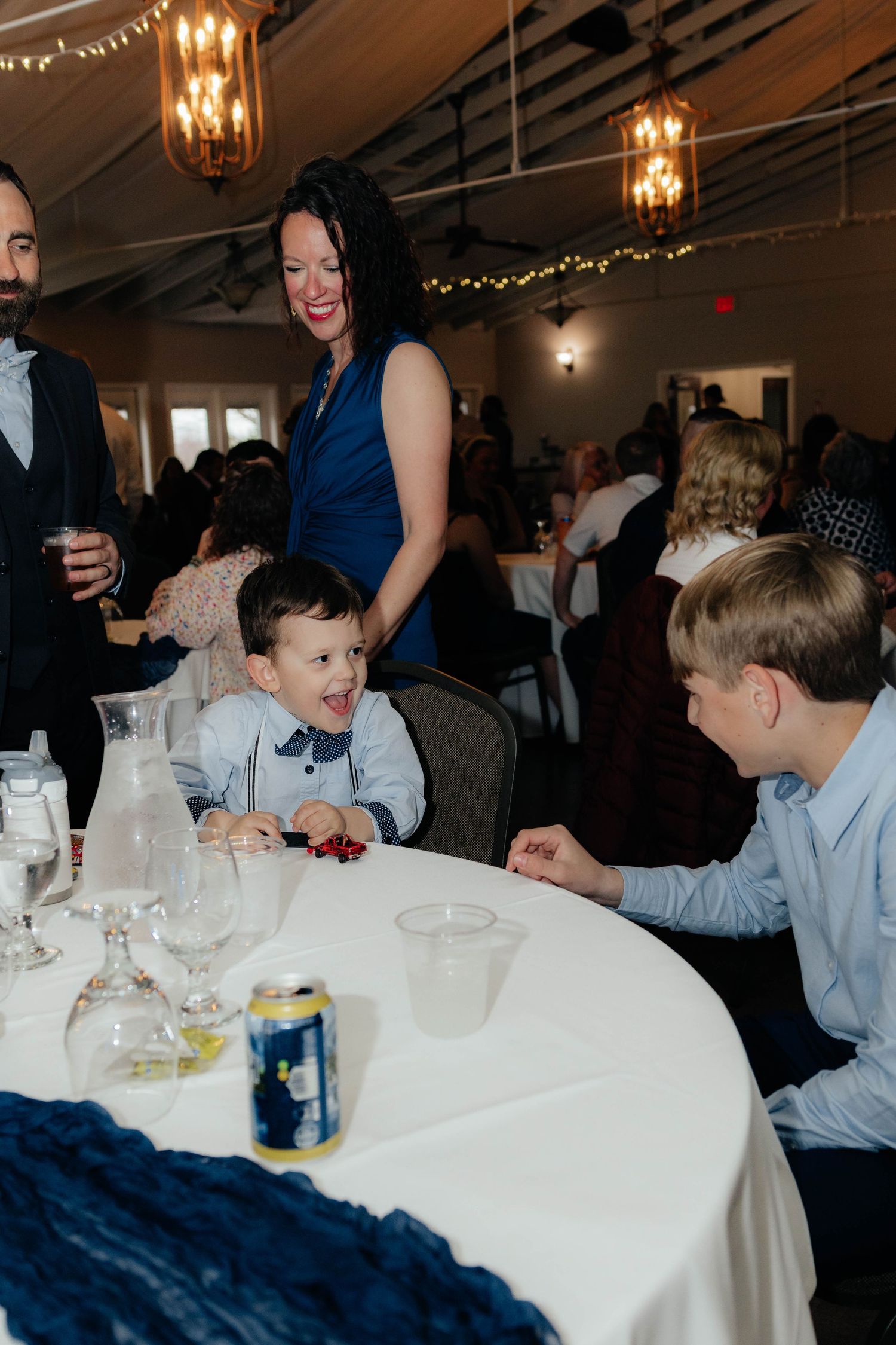 Young boy in suspenders smiles at wedding reception table while woman in blue dress stands nearby.