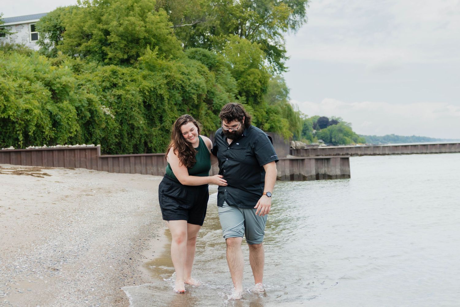 Laughing couple holding hands while wading in shallow lake water during a casual outdoor engagement session.