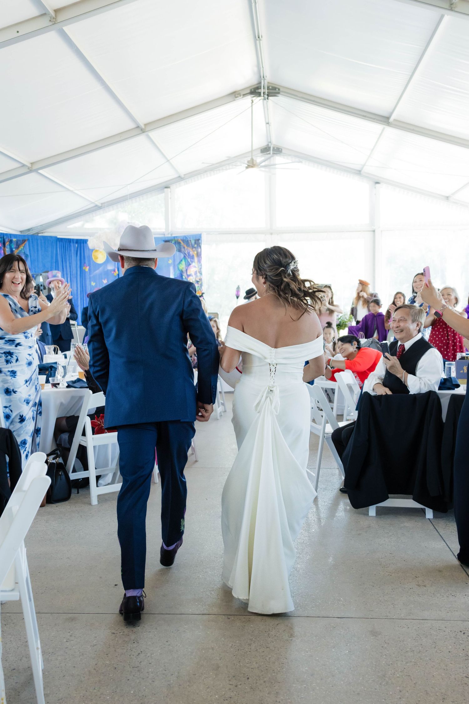 Bride in white off-shoulder gown and groom in navy suit enter wedding reception tent hand in hand.