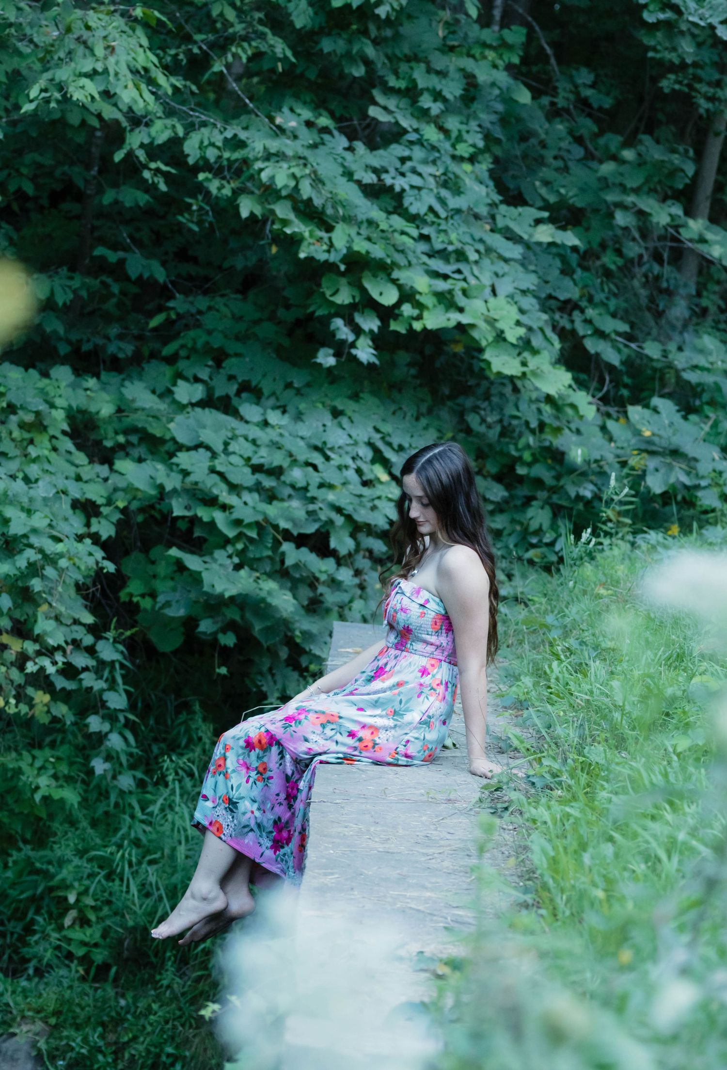 Woman in colorful floral dress sitting on stone path gazing down in serene green woodland setting.