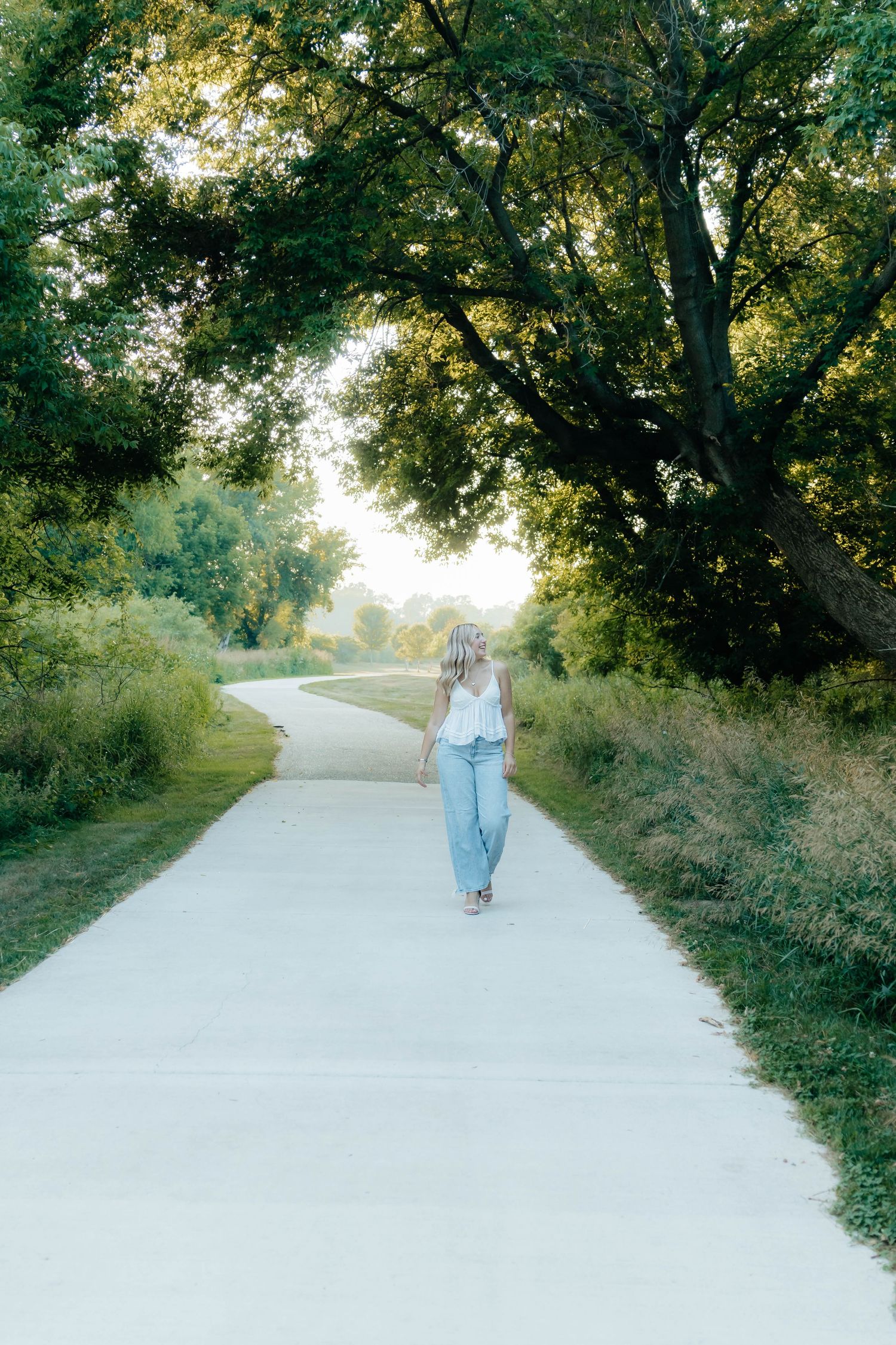 Blonde woman in white top and jeans walking away on scenic tree-lined path at sunset.