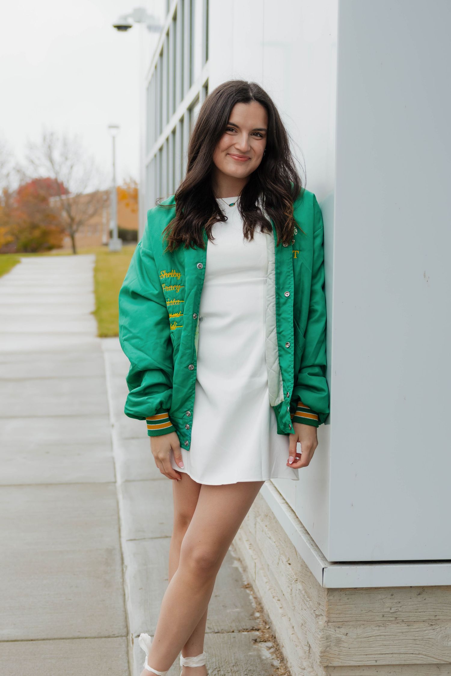 Young woman in a green bomber jacket and white mini dress posing outdoors near a building.