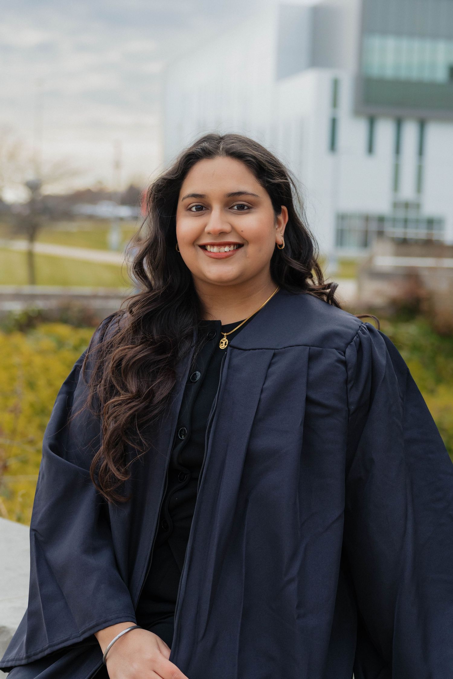 Smiling graduate in navy gown standing outdoors on a university campus on a cloudy day.
