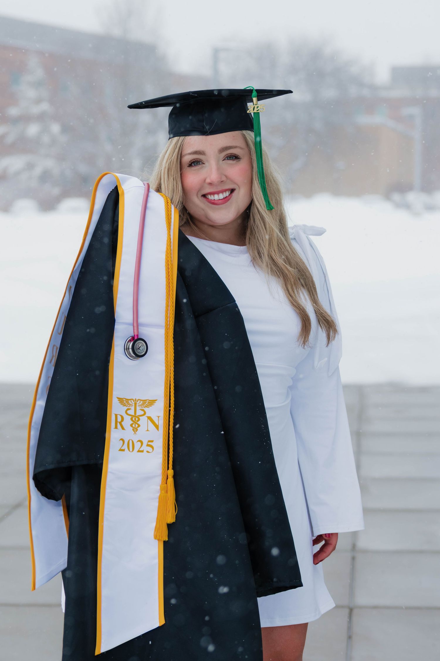 Nursing graduate in cap and gown with RN 2025 stole and stethoscope posing in snowfall.