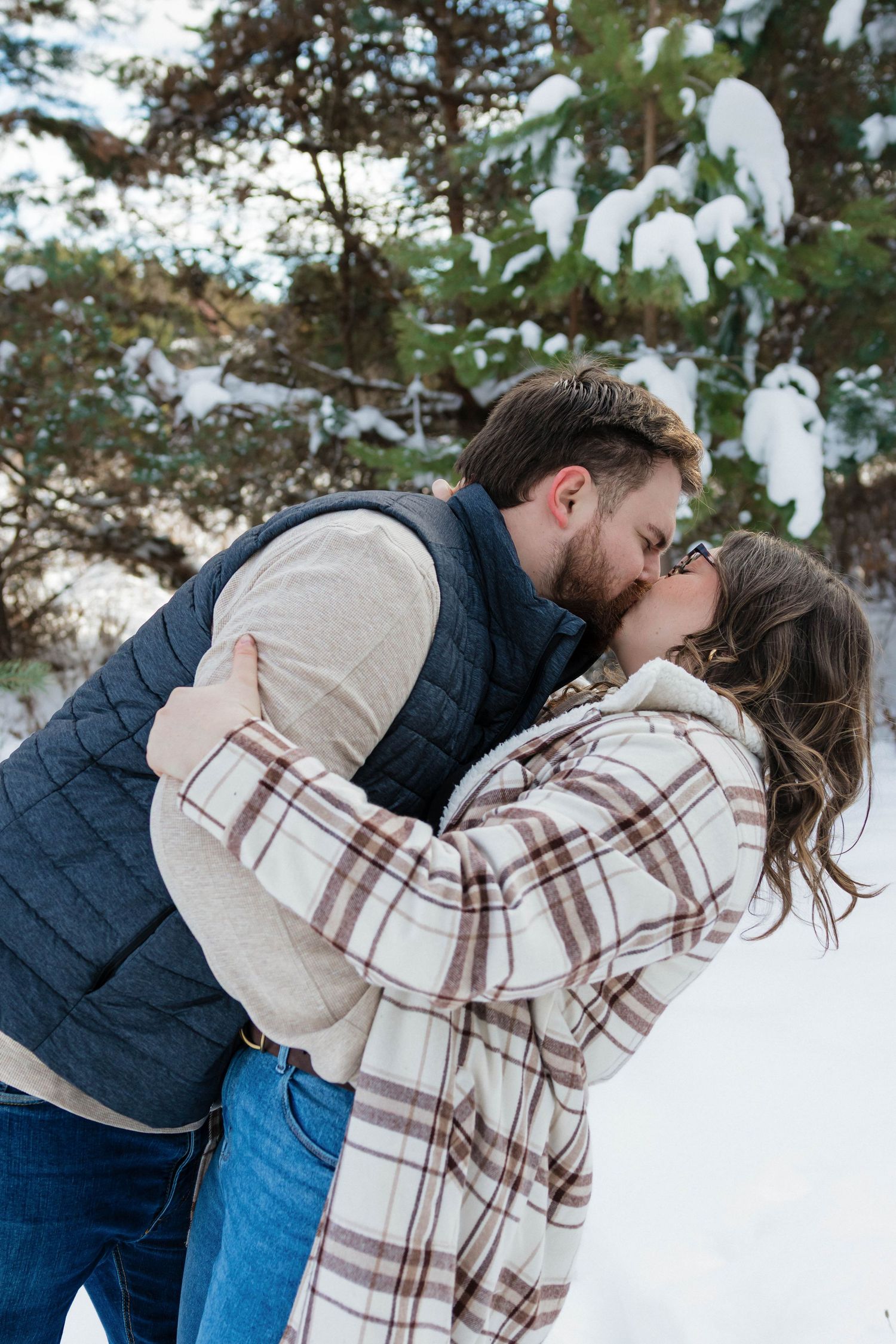 Romantic couple sharing a kiss in a snowy winter forest, woman in plaid jacket and man in quilted vest.