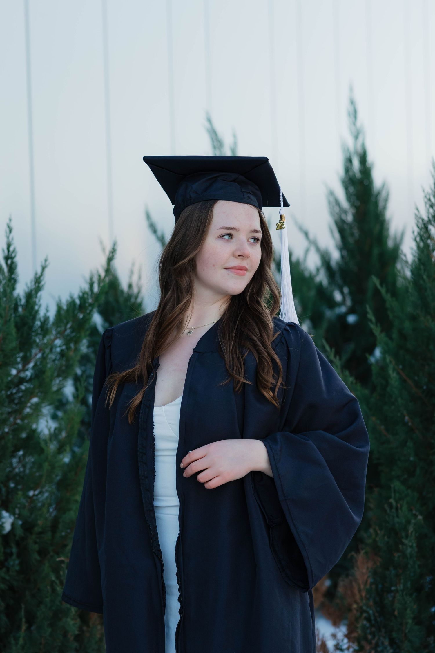 Brunette graduate in black cap and gown gazing thoughtfully beside evergreen trees in winter.