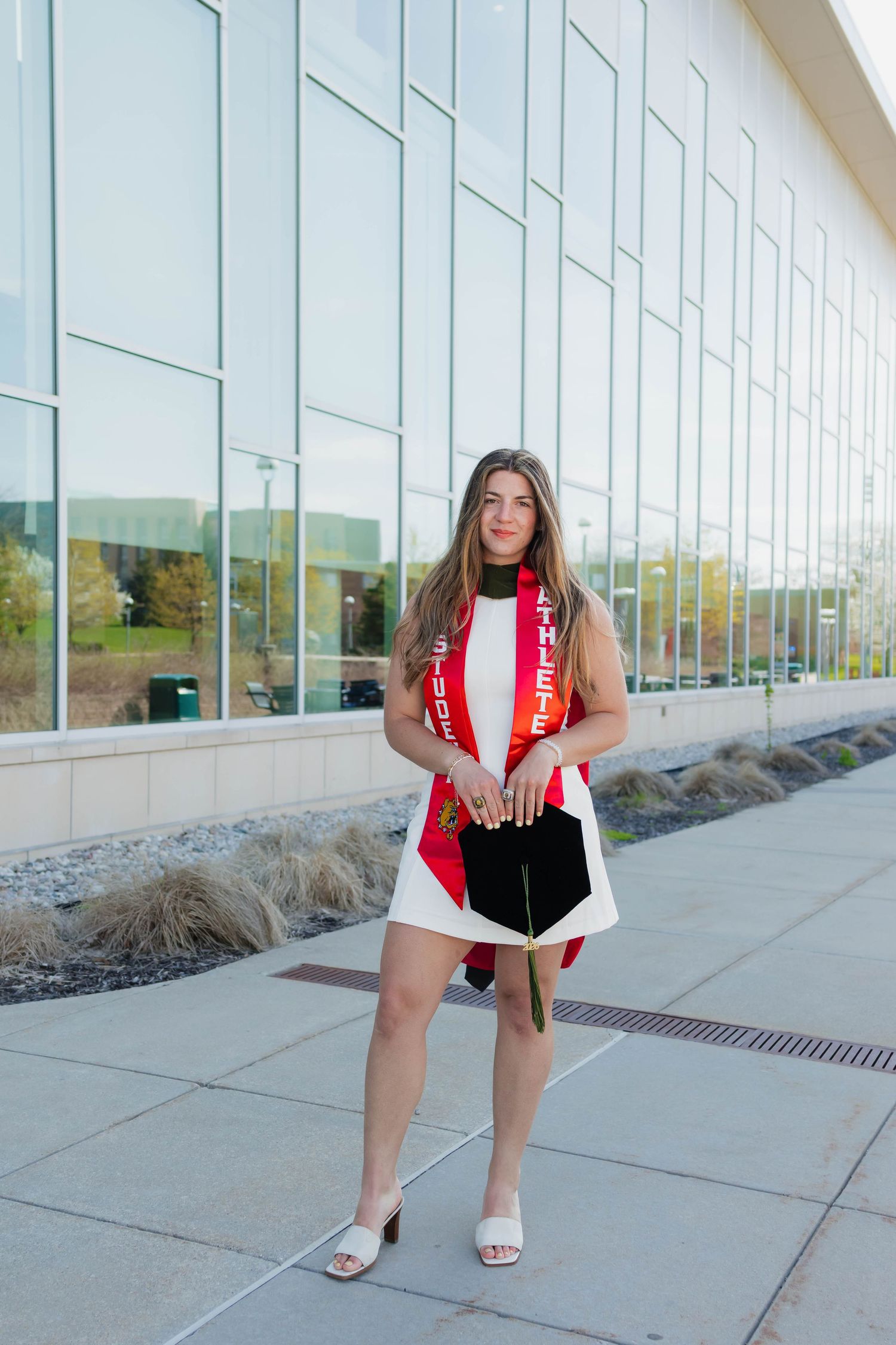 Smiling female graduate wearing red honor stole holding cap outside modern university building.
