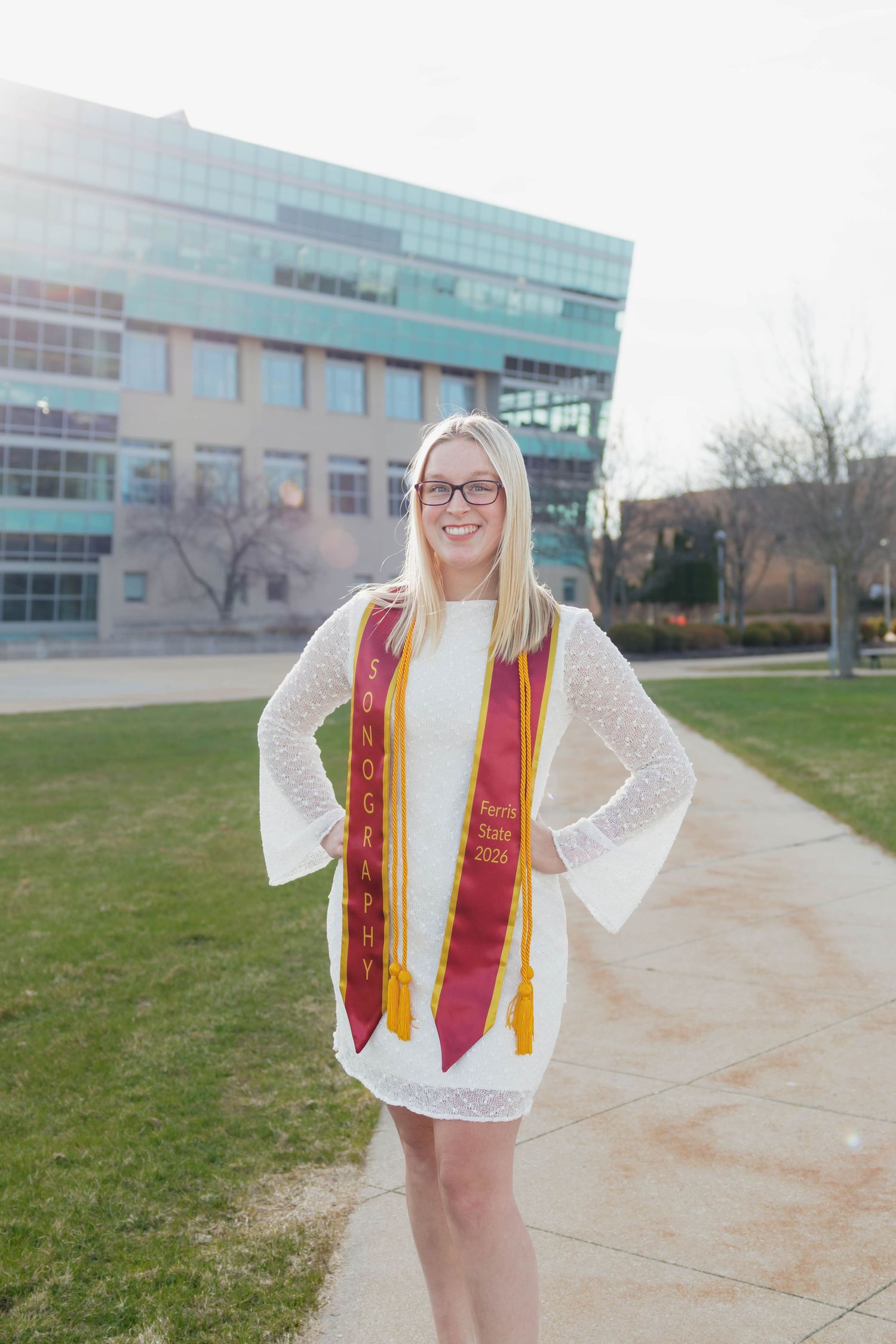 Smiling blonde graduate in white dress wearing a sonography graduation stole posing outdoors on campus.