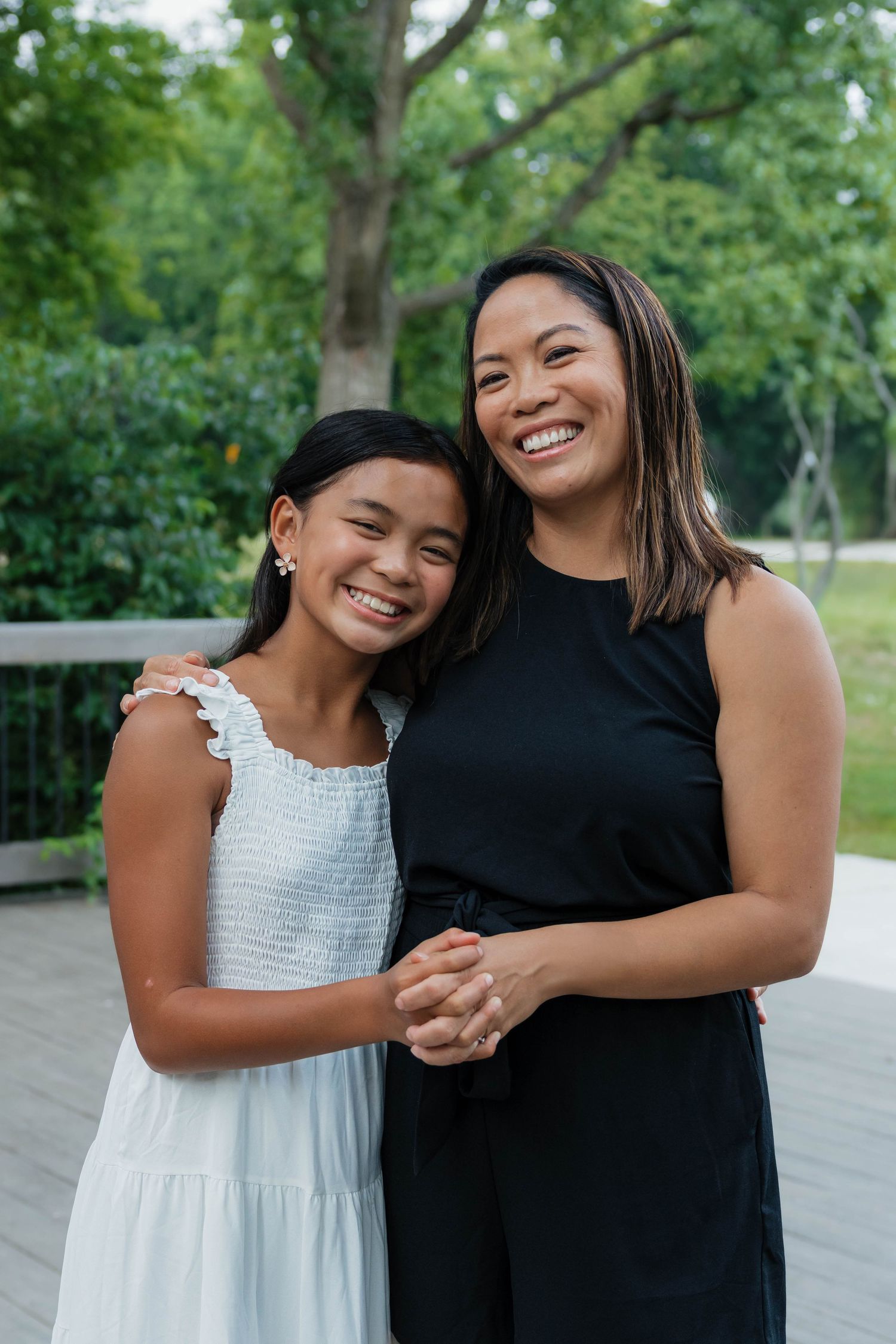 Smiling Asian mother and daughter pose together outdoors in a lush green park setting.