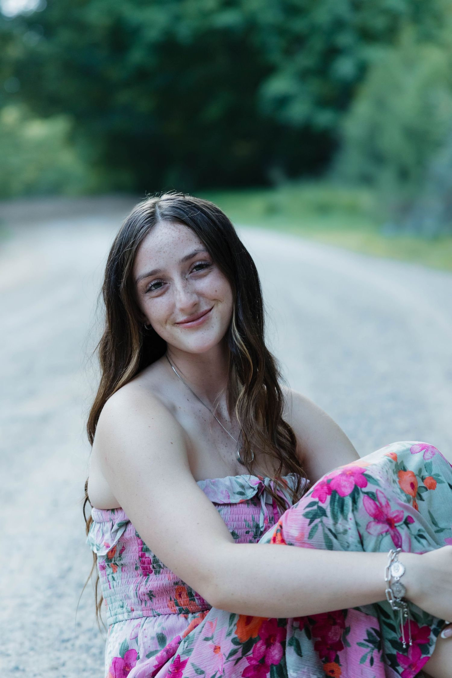 Smiling teenage girl in a floral dress sits on a gravel road with green trees behind her.