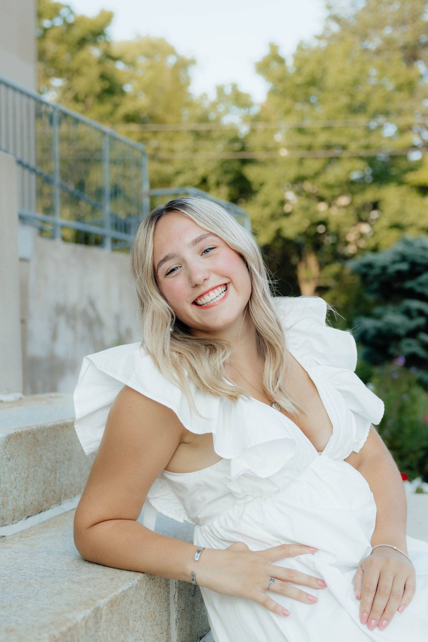 Radiant pregnant woman in white ruffled dress laughing while seated on stone steps outside.