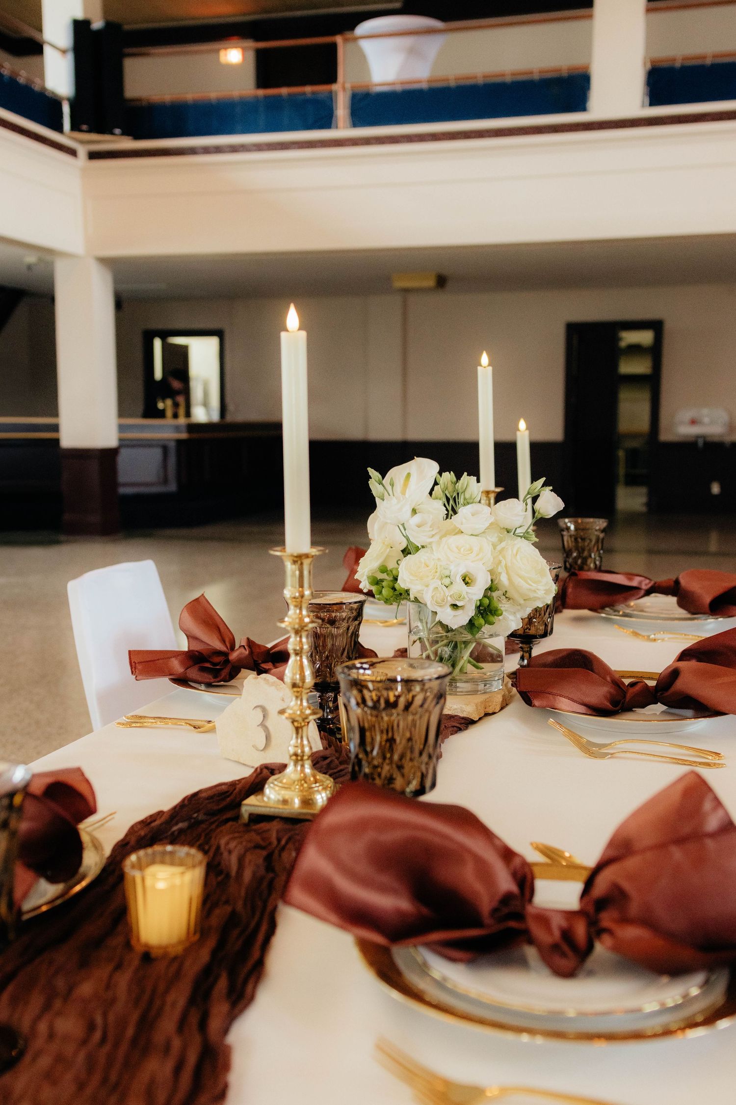 Elegant wedding reception table with burgundy napkins, gold candlesticks, and white floral centerpiece.