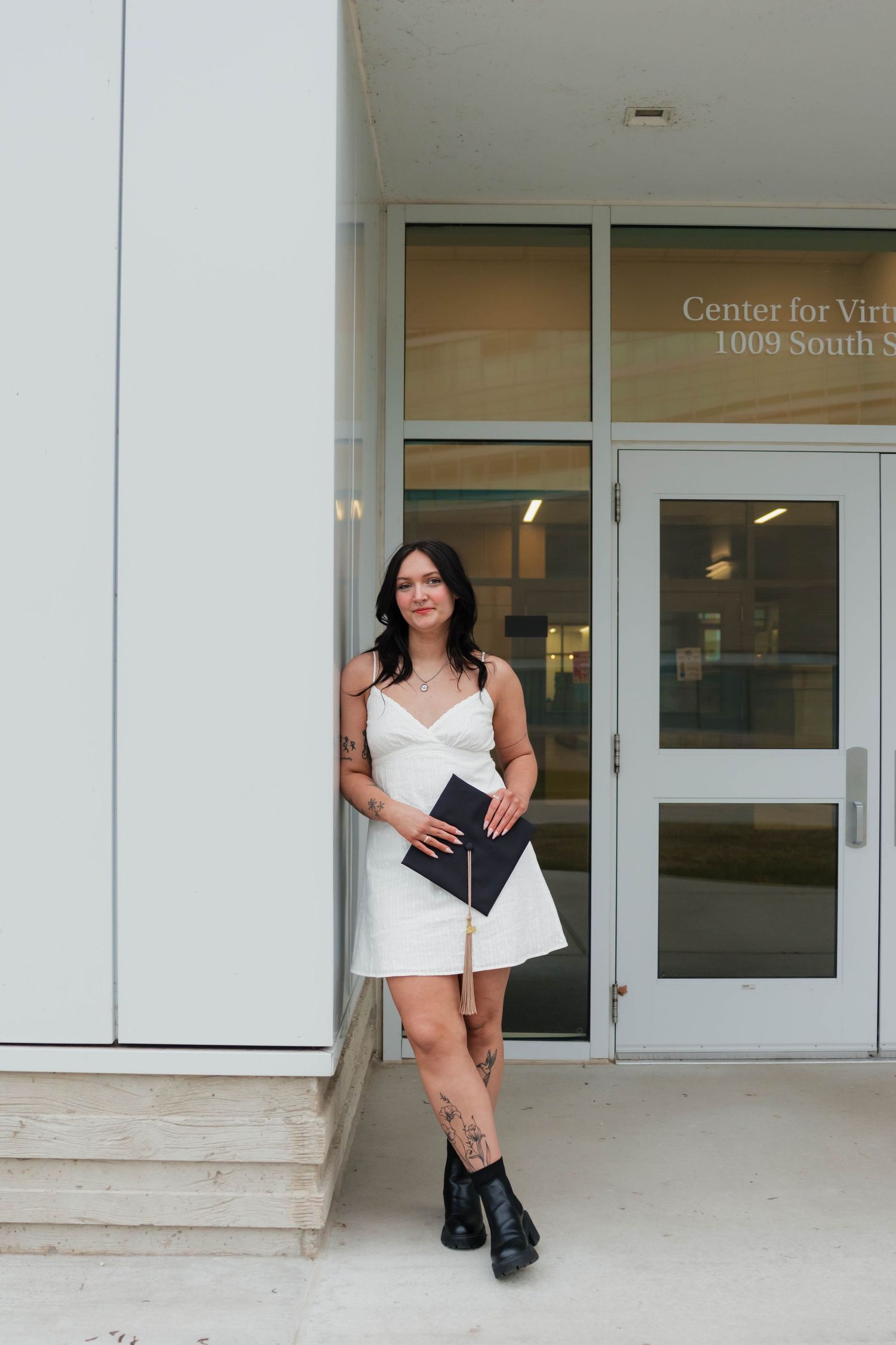 Tattooed brunette graduate in white dress leaning against wall outside Center for Vir building entrance.