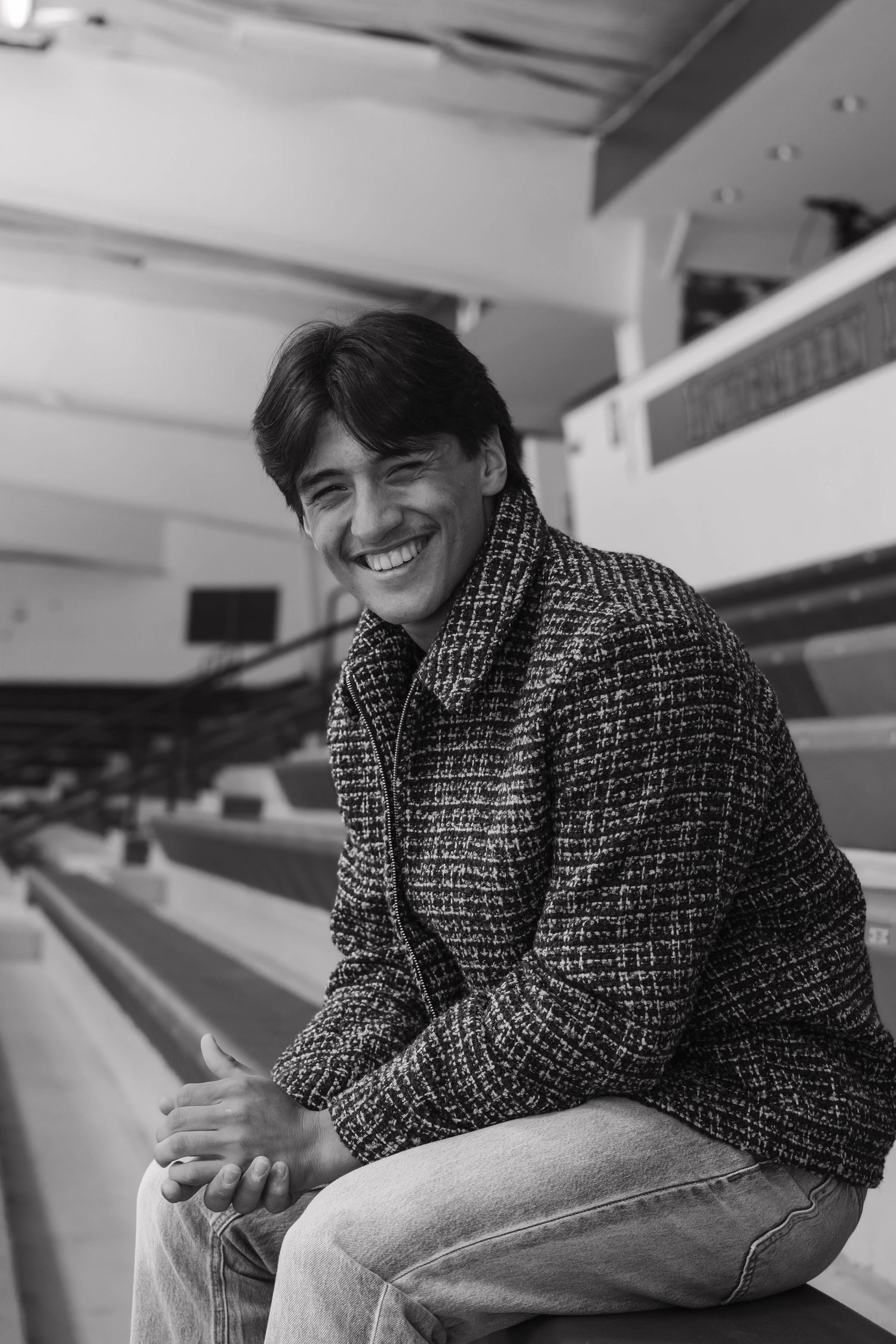 Smiling young man in tweed jacket sits in stadium bleachers, black and white photo.