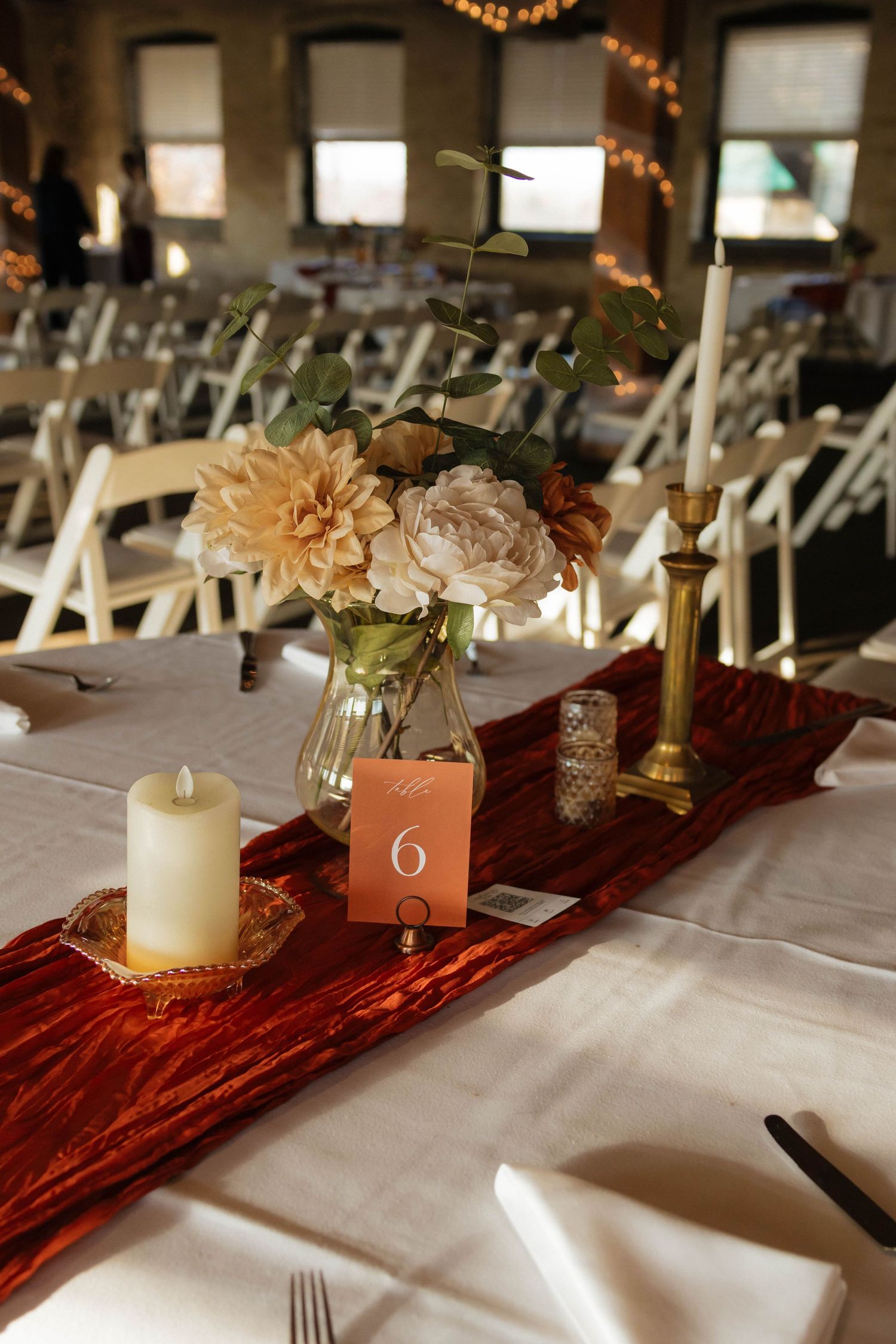 Elegant wedding reception table with red velvet runner, white candle, floral centerpiece, and gold candlestick on white linen.