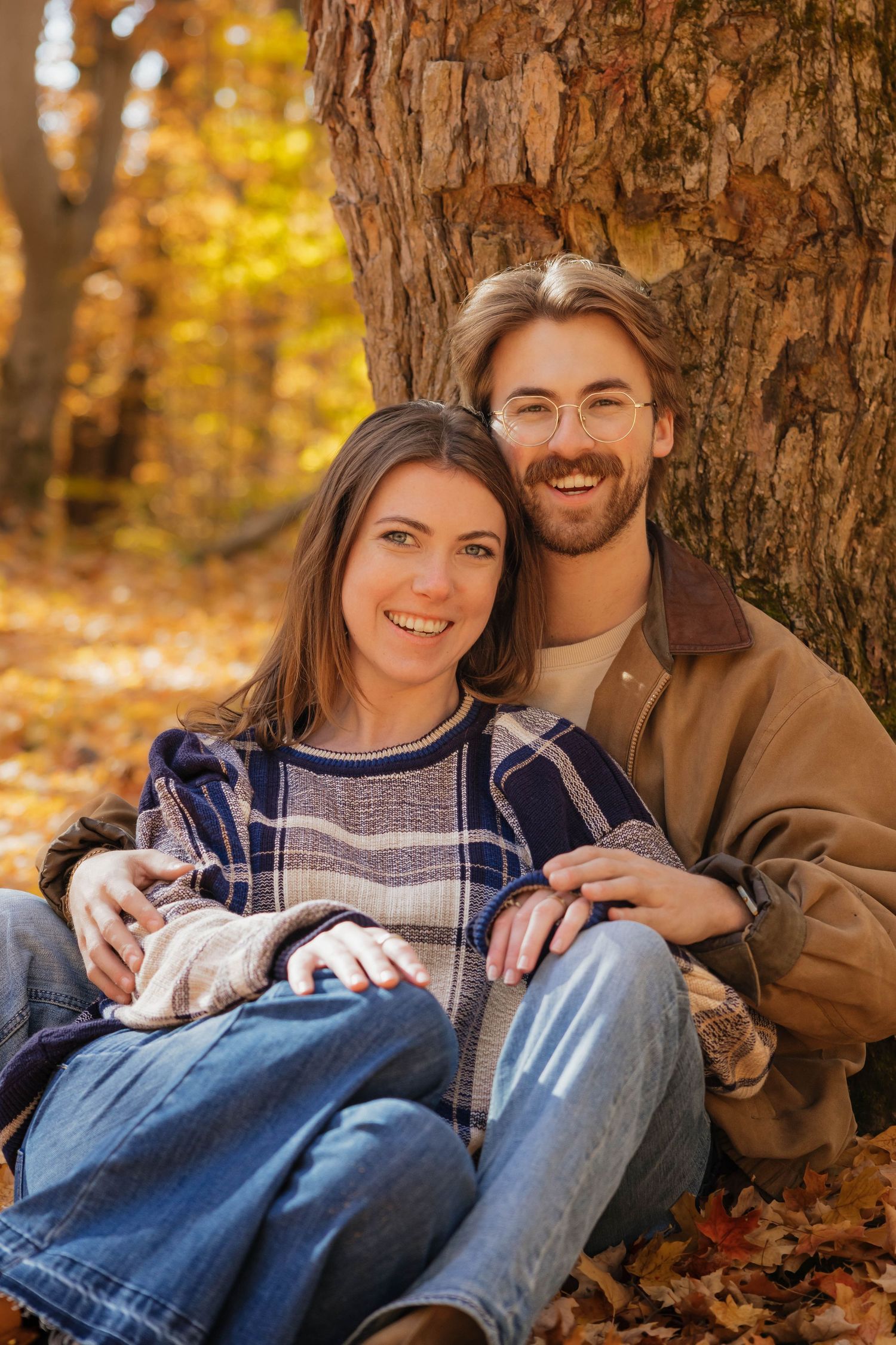 Happy couple smiling and embracing against a tree trunk surrounded by golden autumn foliage.