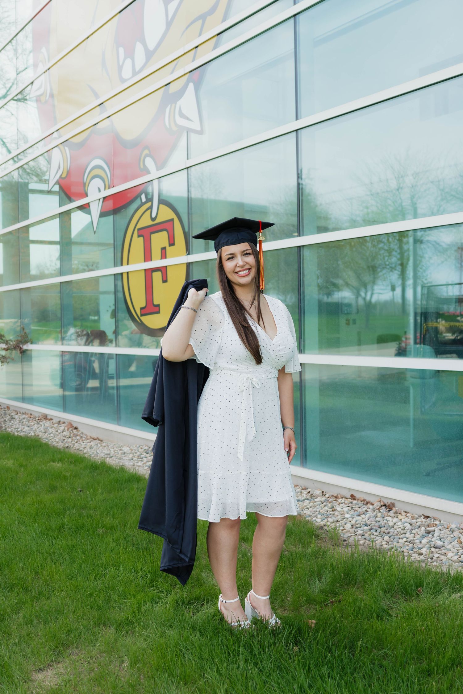 Happy graduate in white dress holds navy gown over shoulder beside building with school logo.