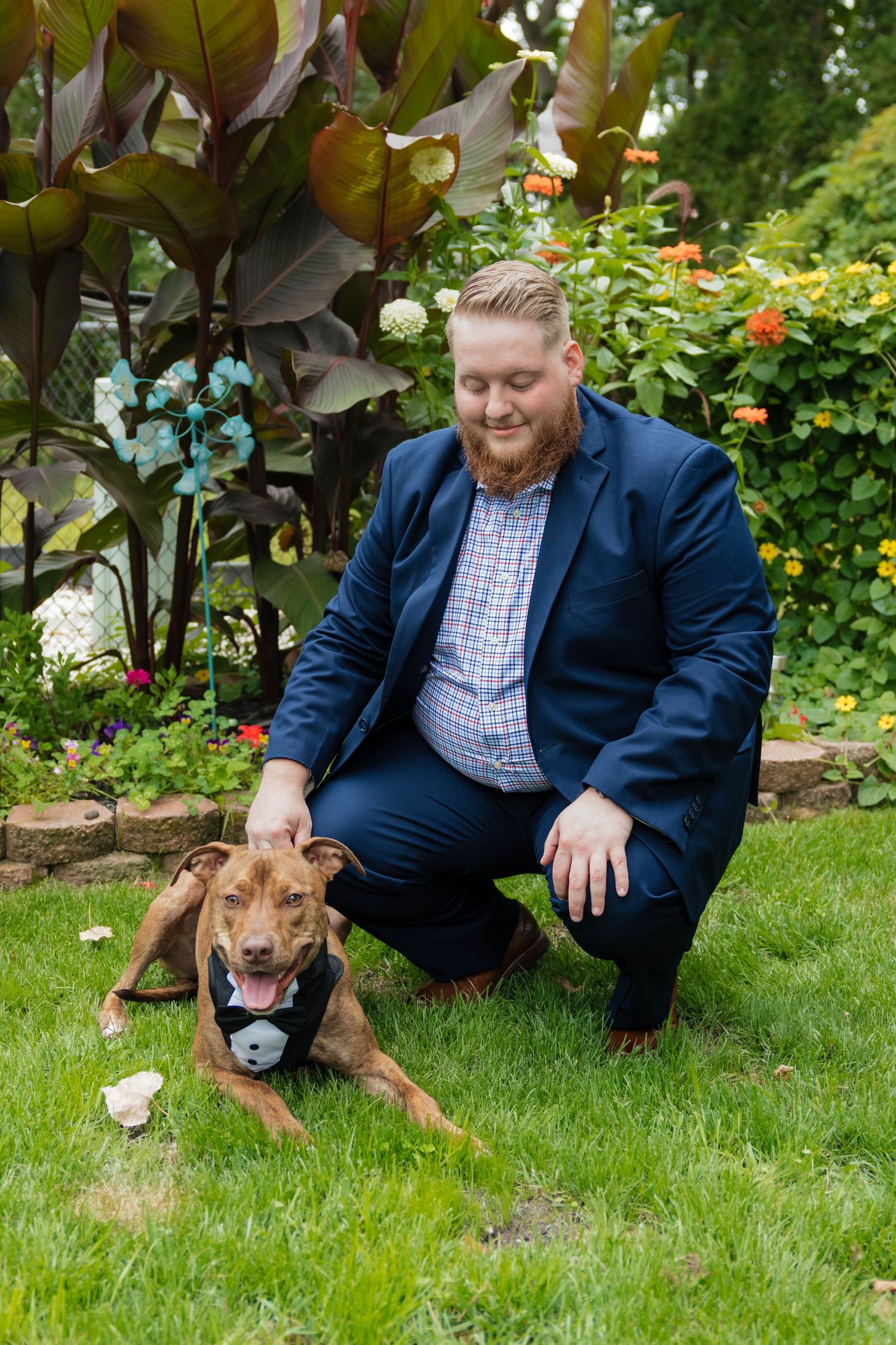 Groom in navy suit petting brown dog in tuxedo bandana in lush garden setting.